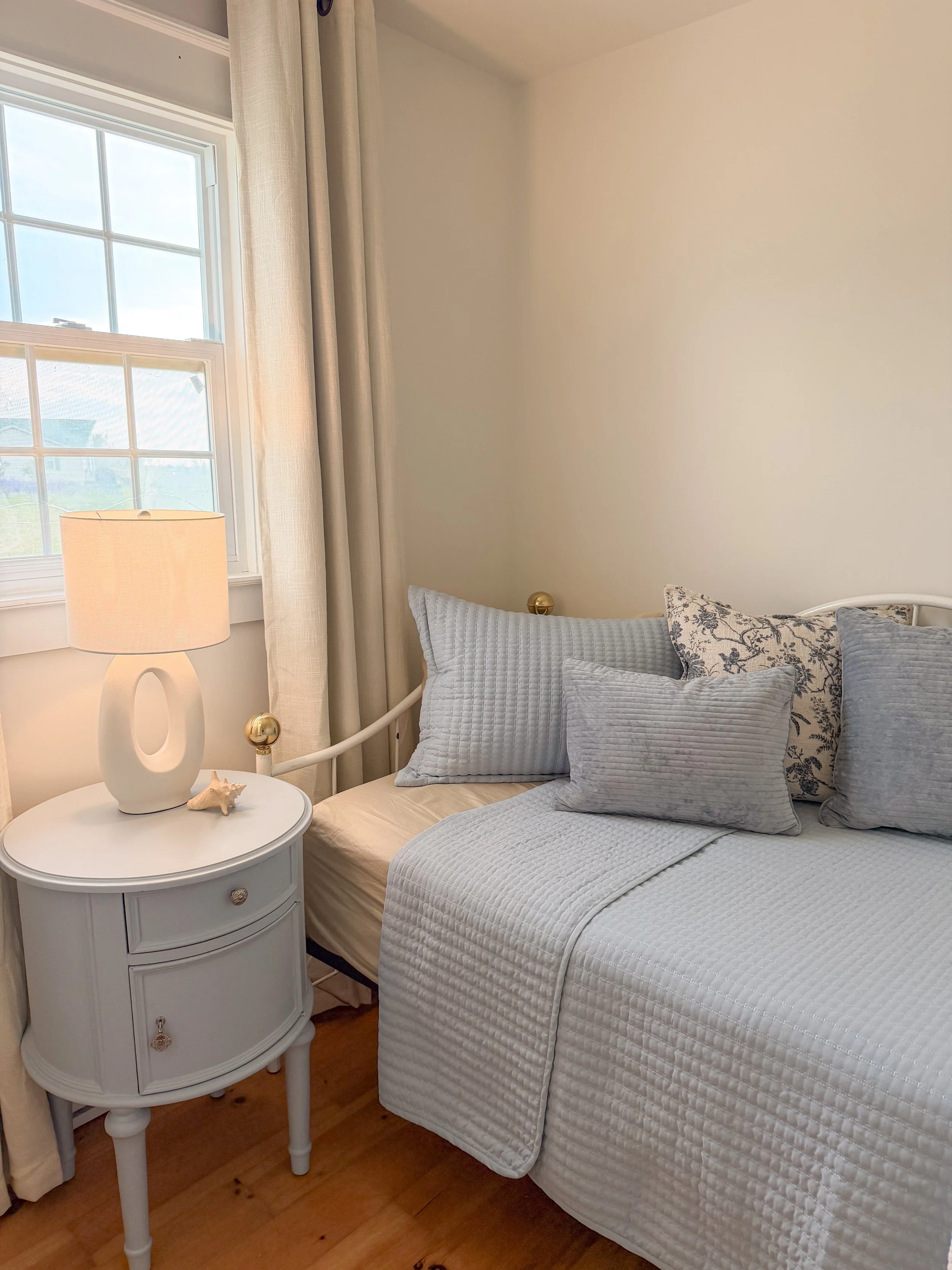 A bedroom corner with a bed covered in white quilted bedding, multiple pillows, a small white bedside table with a single drawer, a cream-colored lamp, a seashell, a window with white trim and divided panes, partially visible cream curtains, and hardwood flooring.