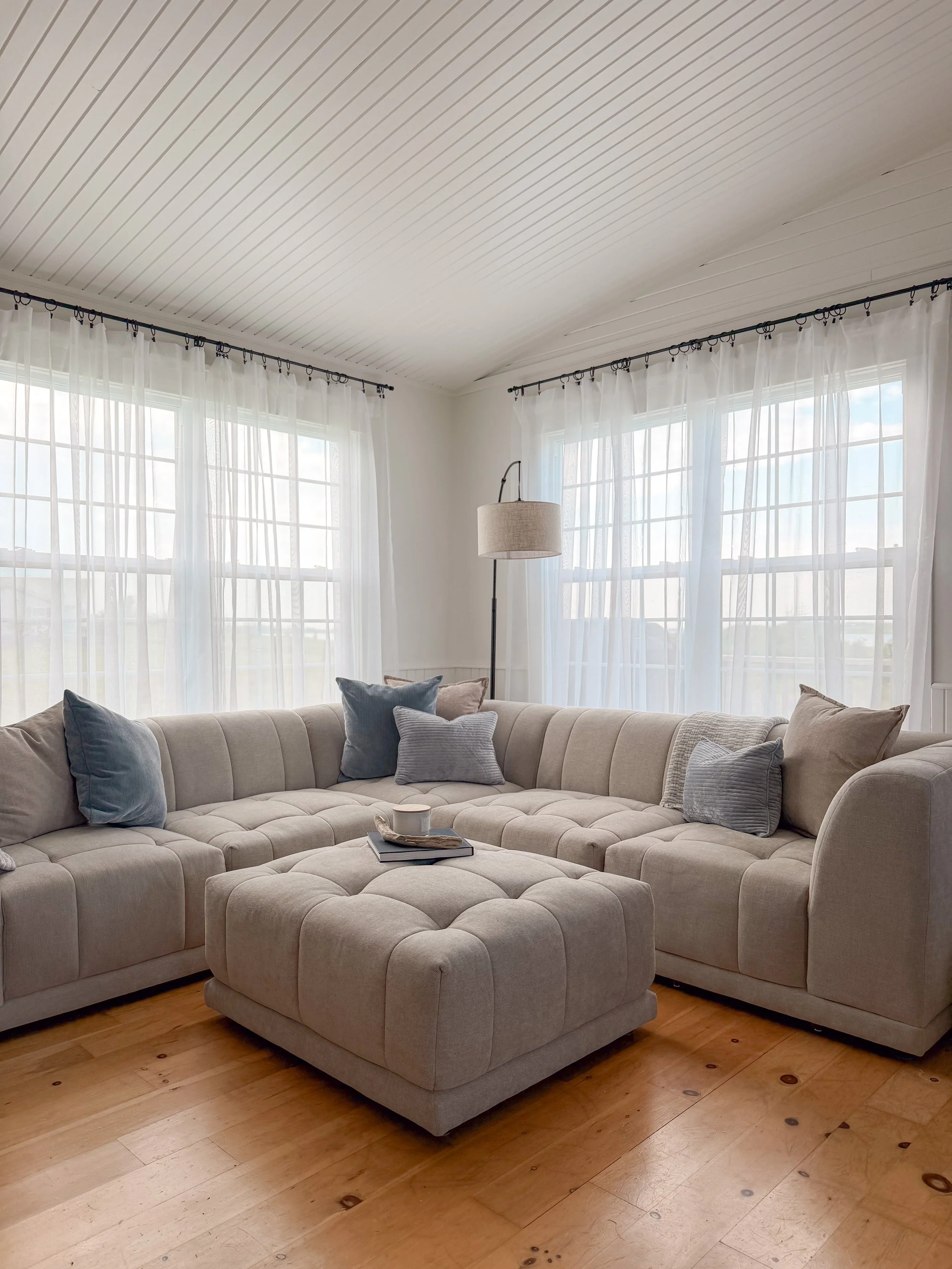 Living room with a beige sectional sofa and matching ottoman, blue and beige pillows, wooden floor, and large windows with sheer white curtains.