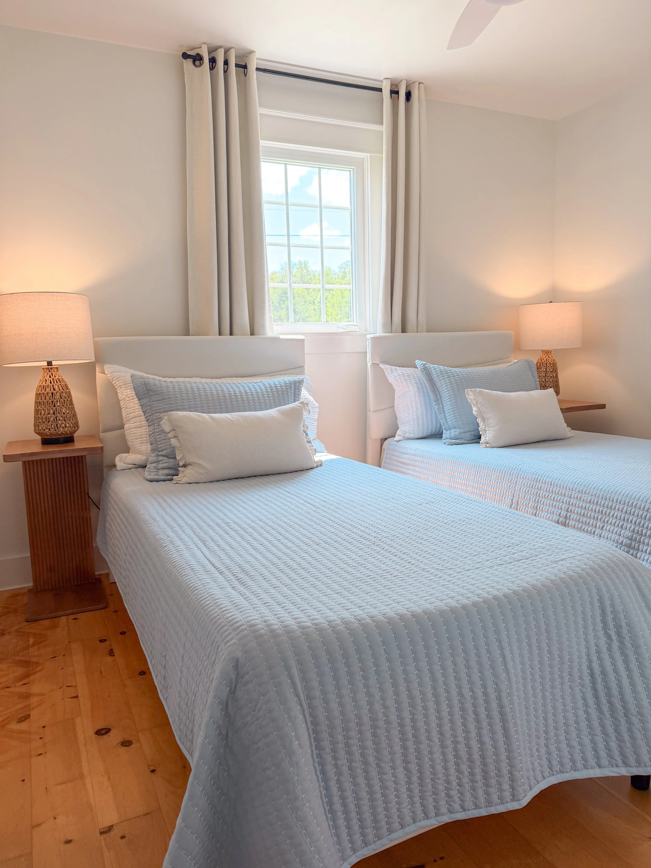A bedroom with two twin beds covered in white quilted bedspreads, each with pillows, and a window with cream curtains between the beds. Two matching lamps sit on wooden bedside tables, with a ceiling fan above.