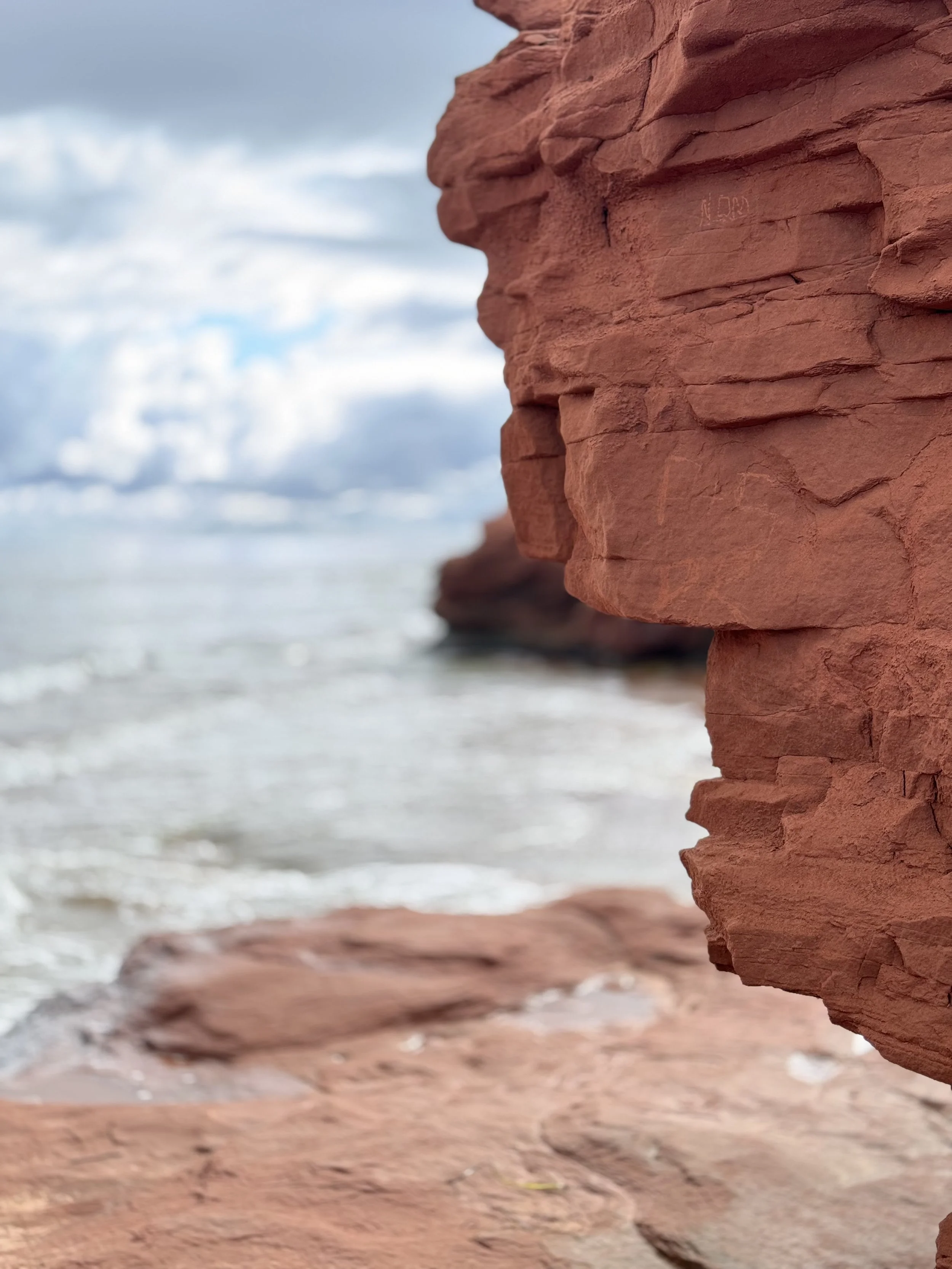 Close-up of a red rocky cliff with a cloudy sky and ocean in the background.