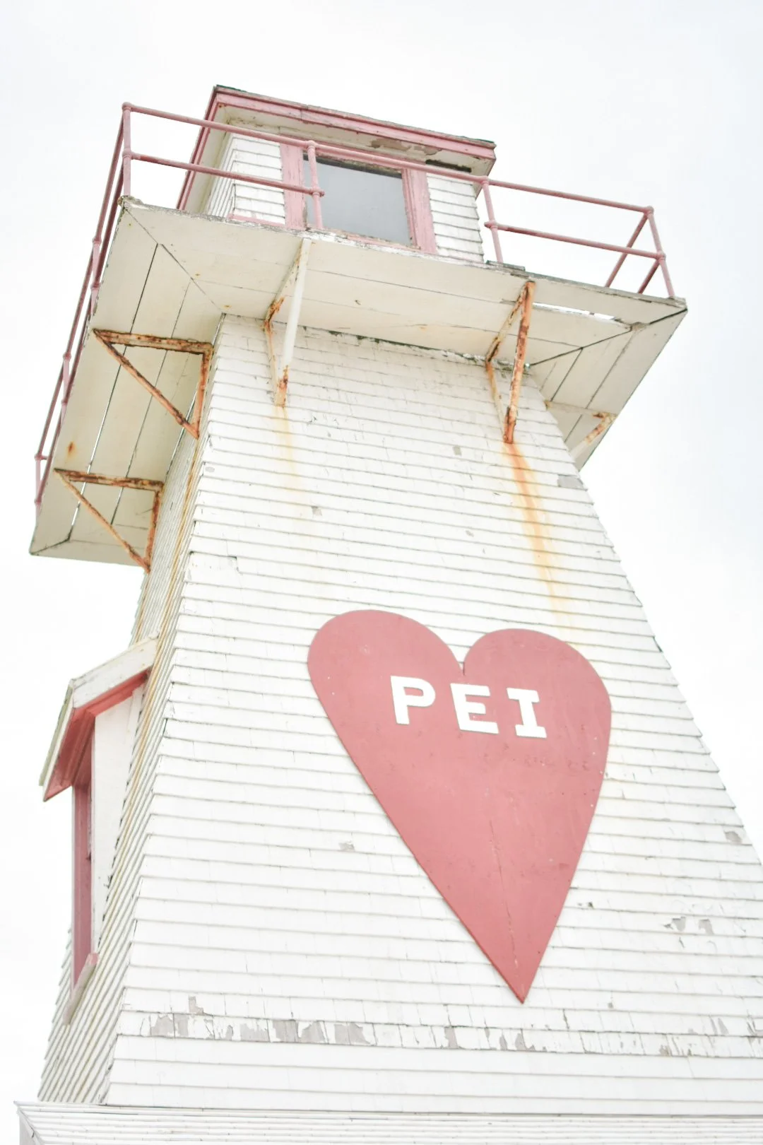 A tall white wooden lighthouse with a pink heart and the word 'PEI' painted inside it, showing signs of rust and weathering, against a cloudy sky.