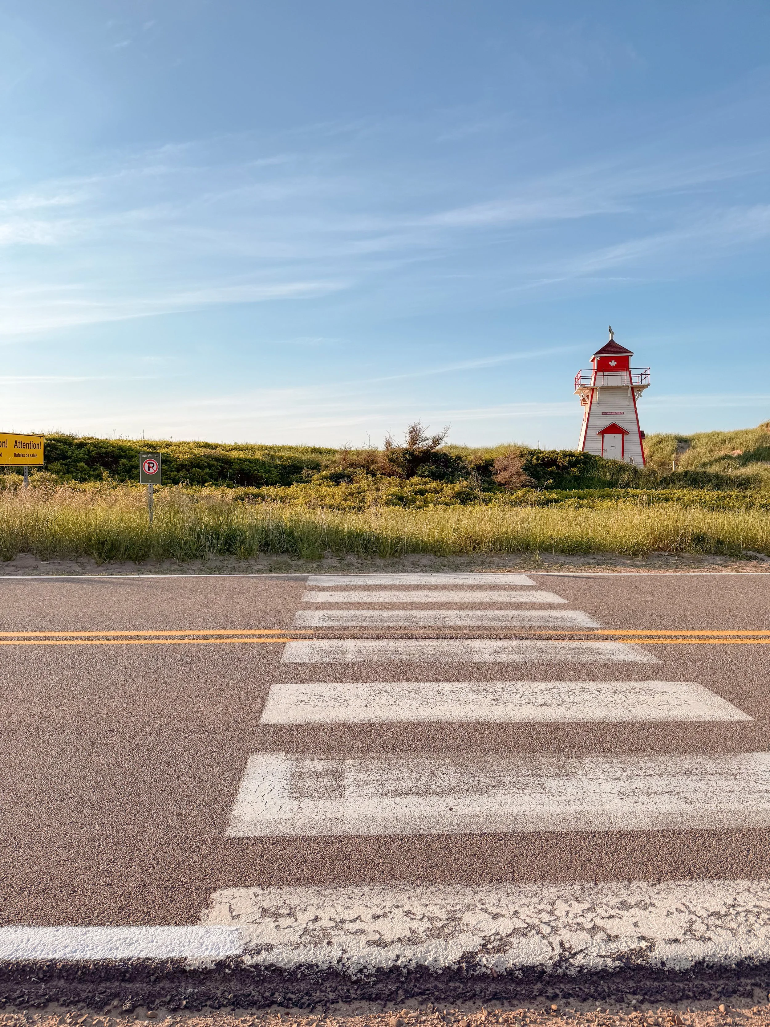 A crosswalk on a road leading to a grassy area with a red and white lighthouse in the background under a blue sky with wispy clouds.
