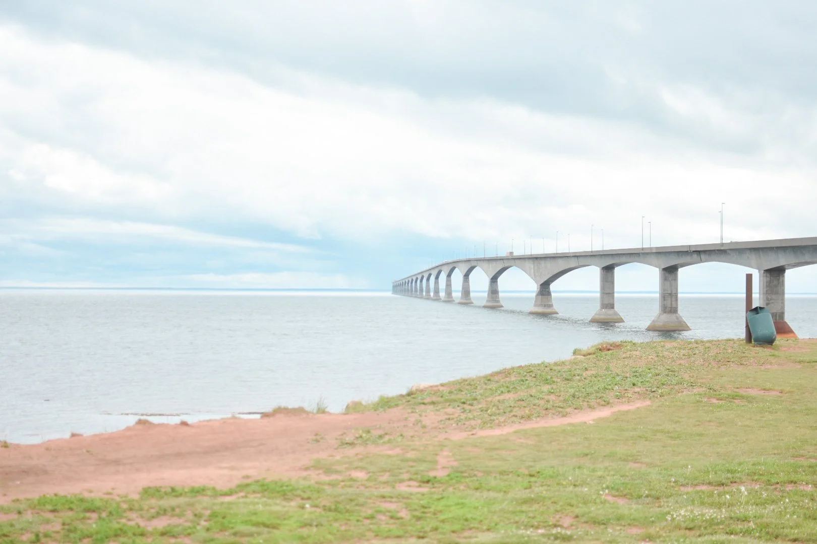 A concrete bridge extends over a body of water with a grassy shoreline in the foreground and cloudy sky above.