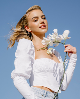 A young woman with long hair stands outdoors against a clear blue sky, holding a white orchid flower, wearing a white off-the-shoulder crop top and light denim shorts.