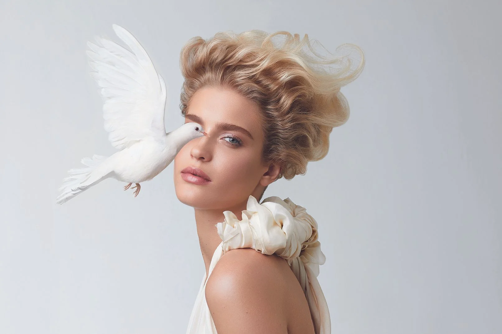 A woman with blonde, wavy hair and a white dress with flower details on the shoulder, posing with a white dove on her face, against a plain light background.