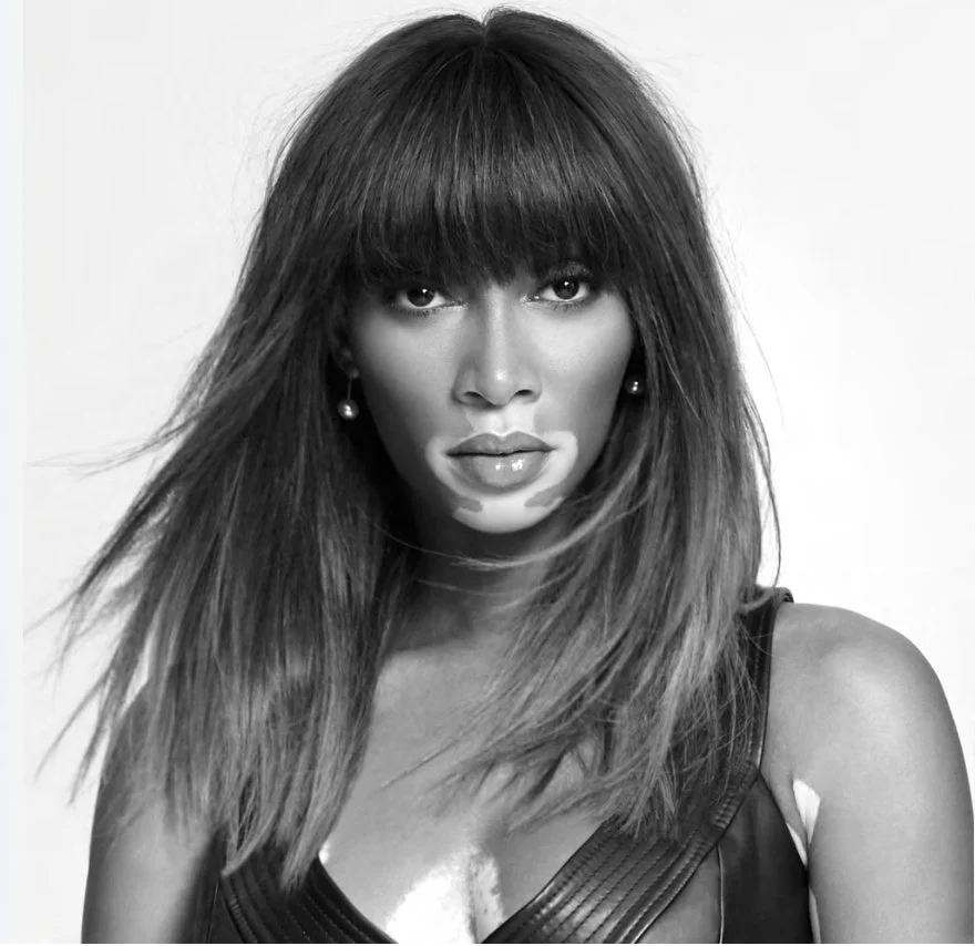 A woman with long hair and bangs, wearing earrings and a leather top, looking directly at the camera in a black-and-white studio portrait.