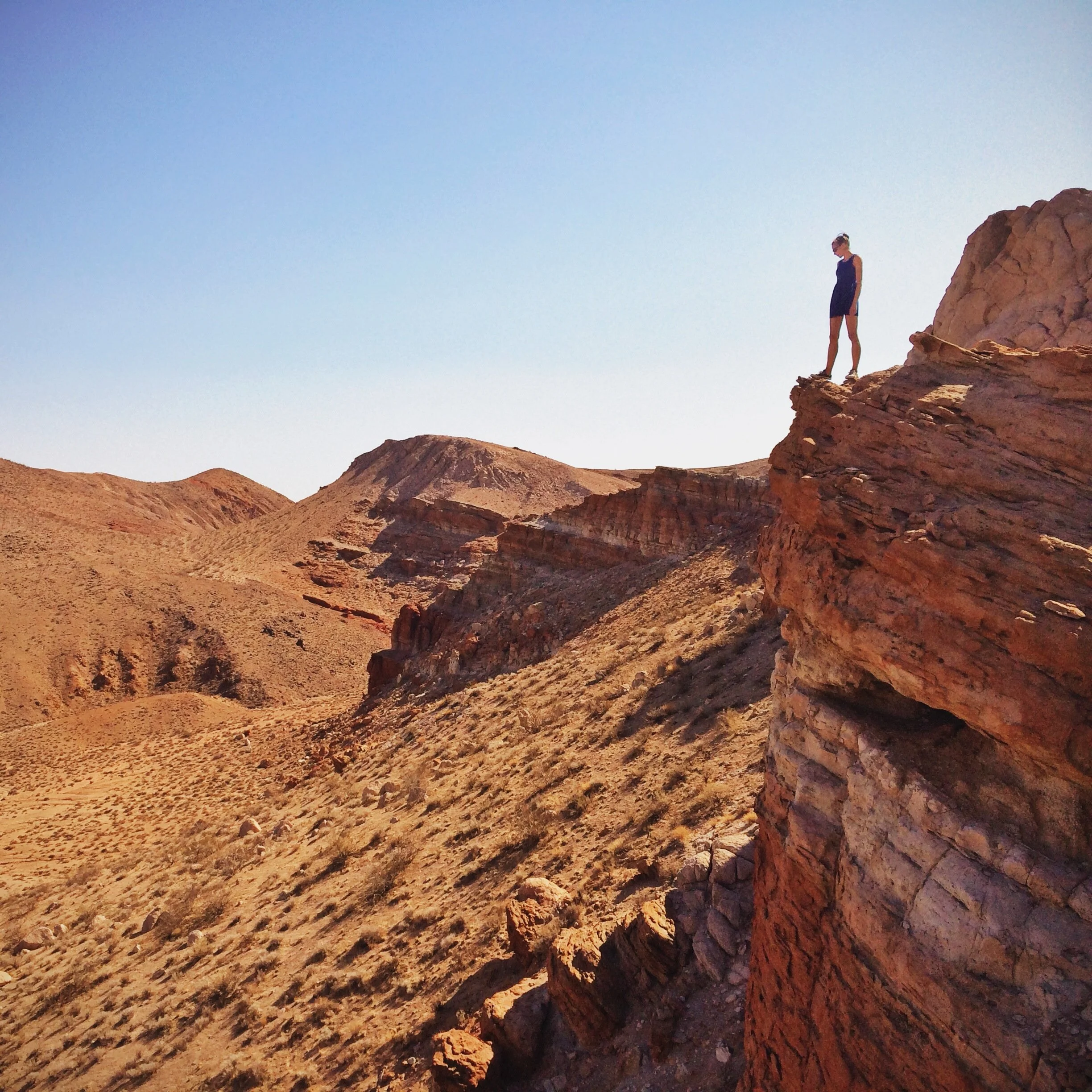 A woman in a black dress standing on the edge of a rocky cliff under a clear blue sky in a desert landscape.