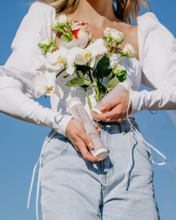 Person holding a bouquet of white and green flowers, wearing a white long-sleeve top and light blue jeans, against a clear blue sky.