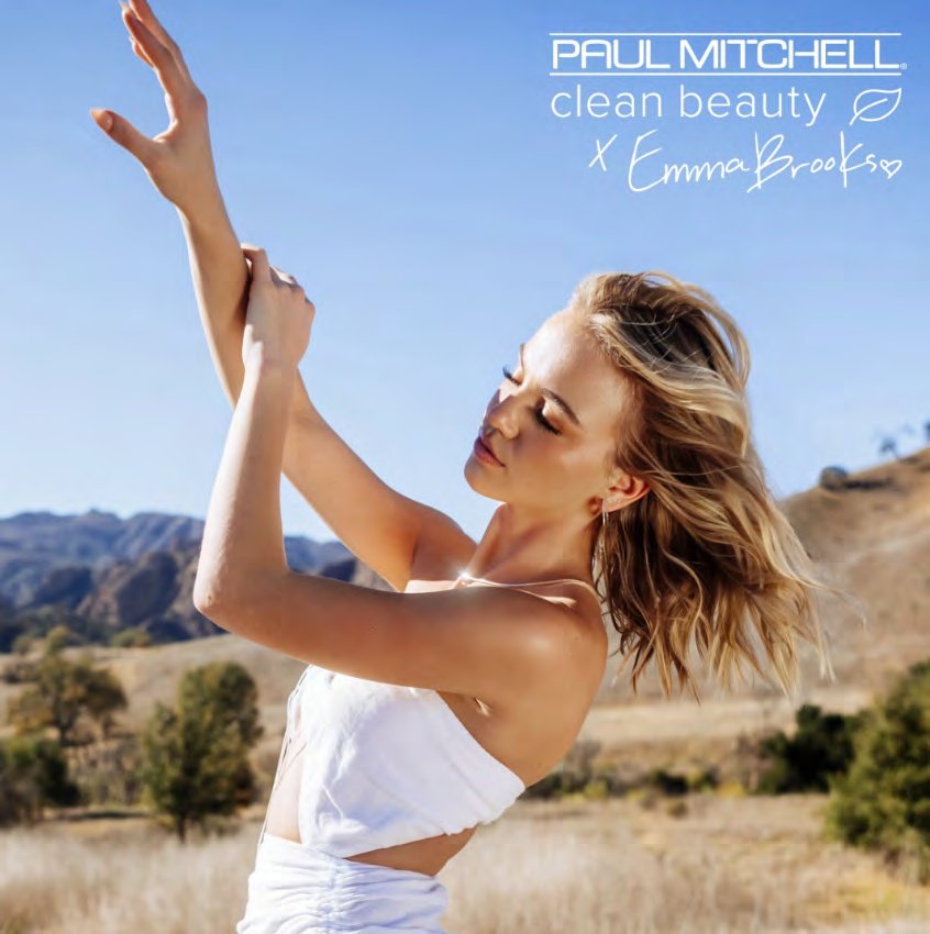 A woman outdoors in a scenic landscape raises her arms with her eyes closed, wearing a white tank top, with mountains and trees in the background, promoting Paul Mitchell's Clean Beauty by Emma Brooks.