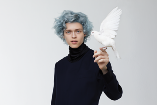 Person with short, curly, gray hair holding a white dove in front of a plain background.