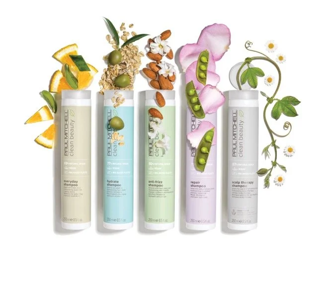 Five bottles of Paul Mitchell hair care shampoos with herbs and fruits around them, including oranges, olives, almonds, pink flower petals, and green peas, on a white background.