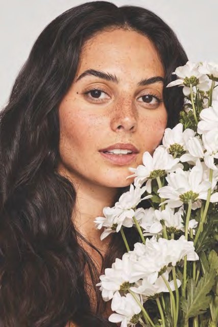 Close-up of a young woman with long dark hair and freckles, holding a large bouquet of white flowers close to her face.