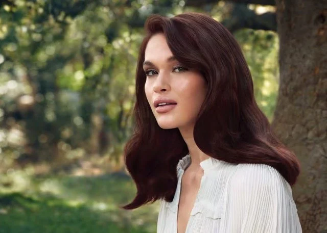 A woman with long, wavy auburn hair standing outdoors near a tree in a park, wearing a white blouse, with a blurred green background.