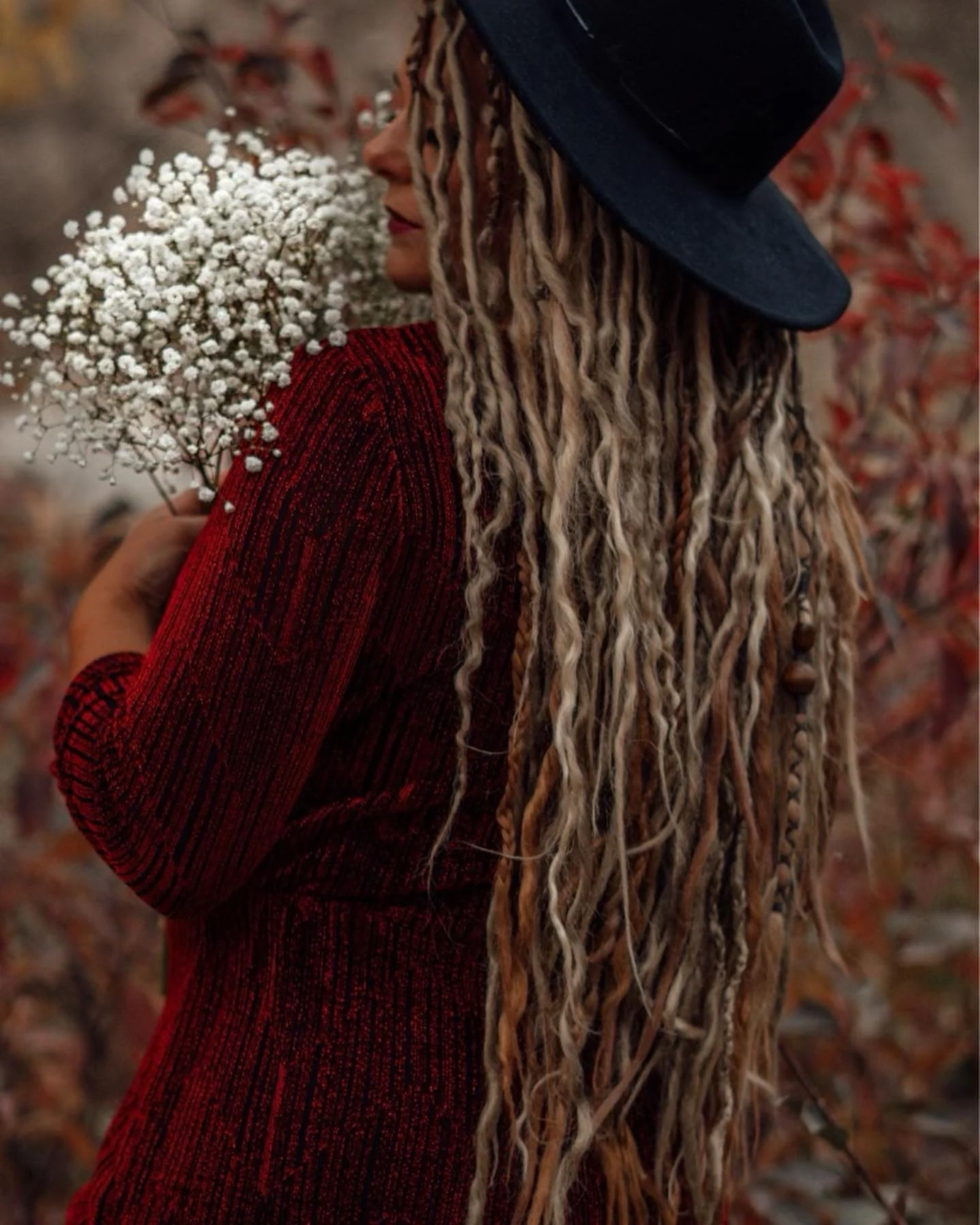 A woman with long dreadlocks, wearing a wide-brimmed hat and red textured sweater, holding a bouquet of small white flowers outdoors with autumn-colored foliage in the background.