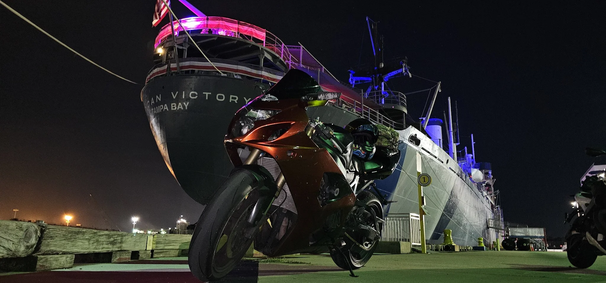 Nighttime scene featuring a large ship named 'San Victory' docked at a port, illuminated with colorful lights. In the foreground, a red motorcycle is parked on a concrete surface.