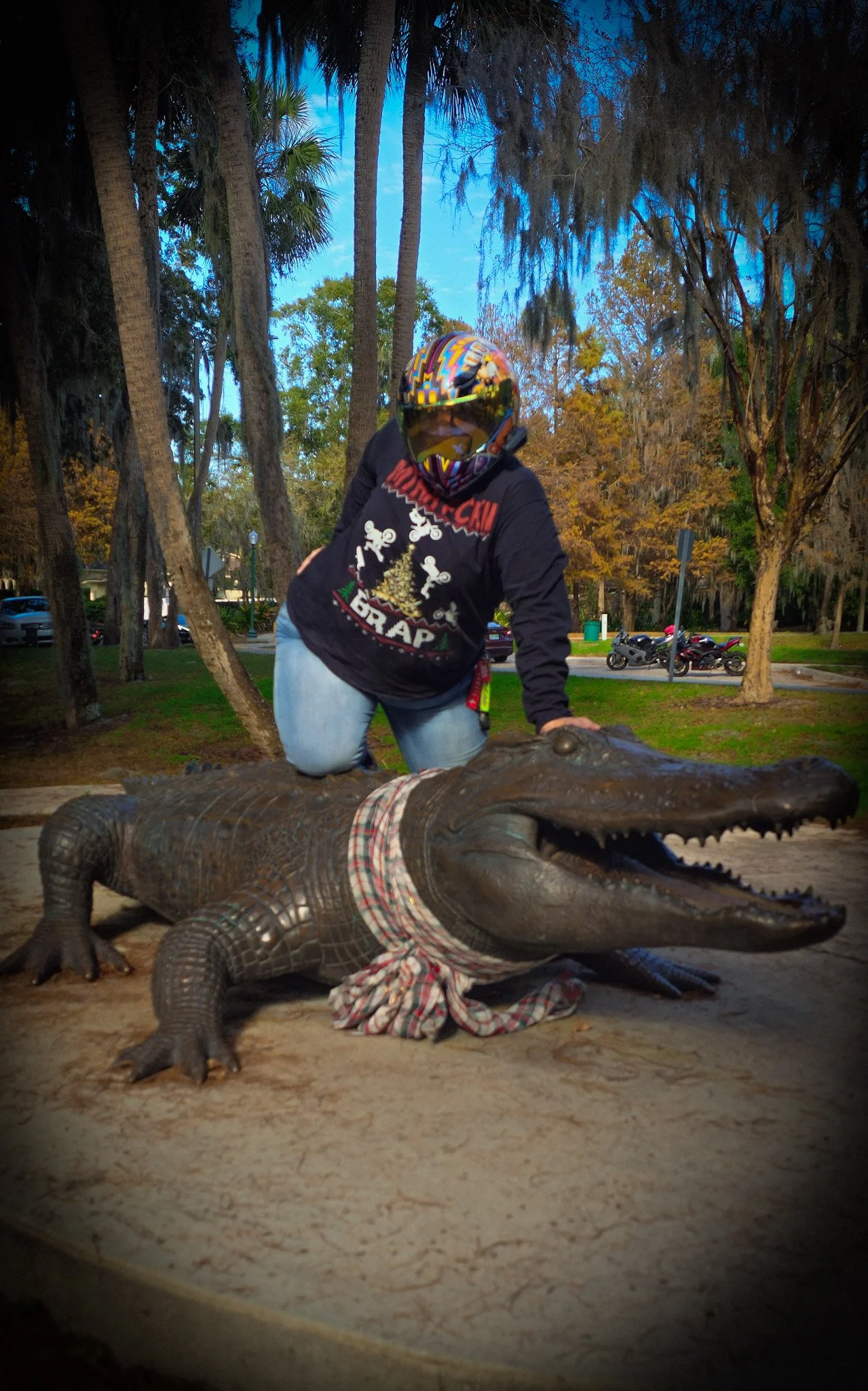 A person wearing a colorful helmet and a black hoodie with graphic designs is kneeling on a large crocodile's back, with the crocodile's mouth open wide, revealing its teeth. The scene is outdoors with trees and parked motorcycles in the background during evening or night.