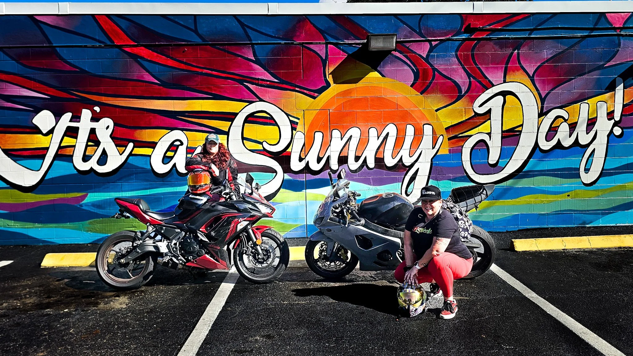 Two women are posing with motorcycles in front of a colorful mural that reads 'It's a Sunny Day!'. One woman is wearing a black jacket and pink pants, and the other is wearing a black T-shirt and blue jeans, both smiling and holding motorcycle helmets.