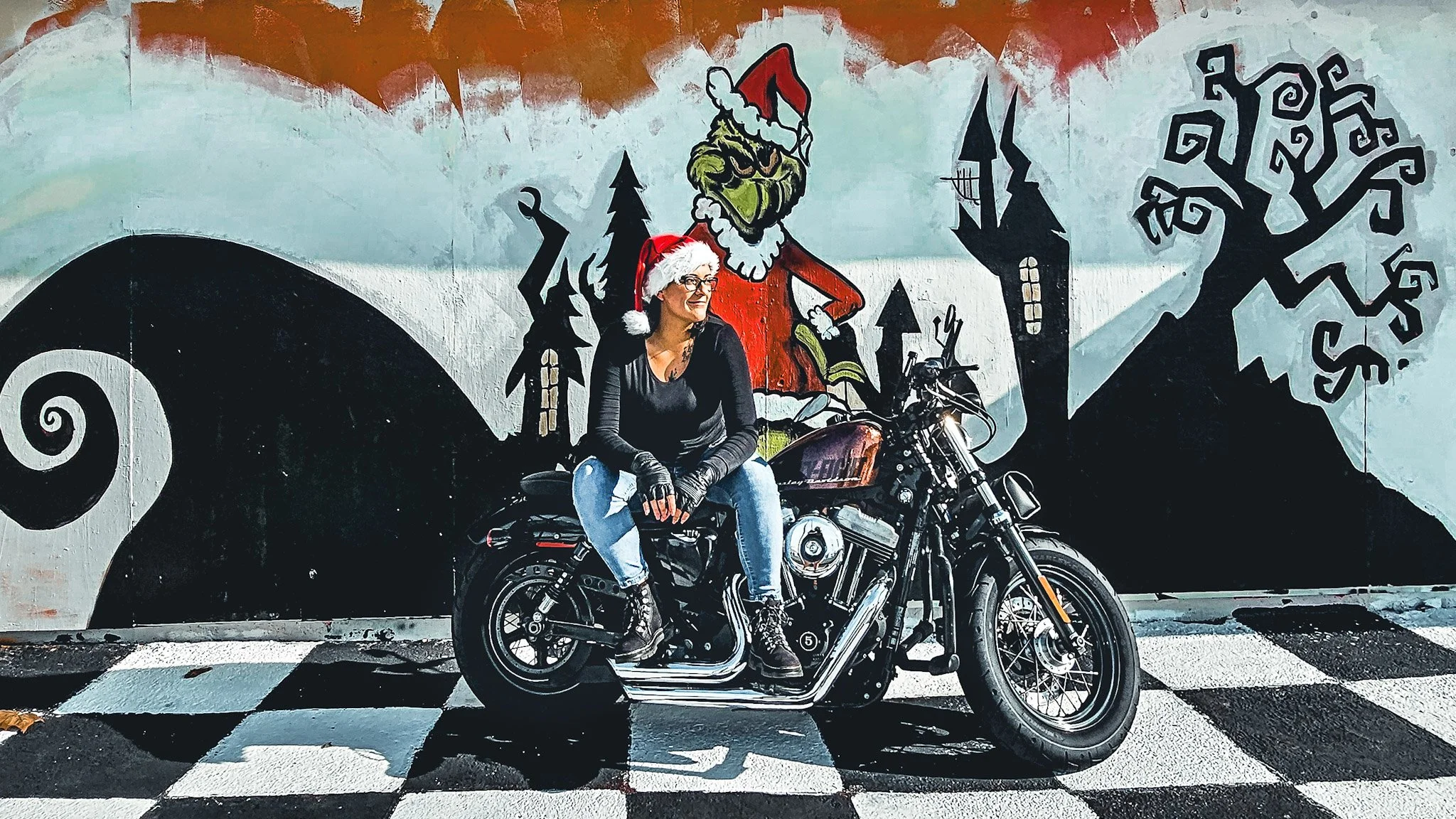 Woman with a Santa hat sitting on a black motorcycle in front of a mural featuring a grinch in a Santa hat, trees, and a haunted house.