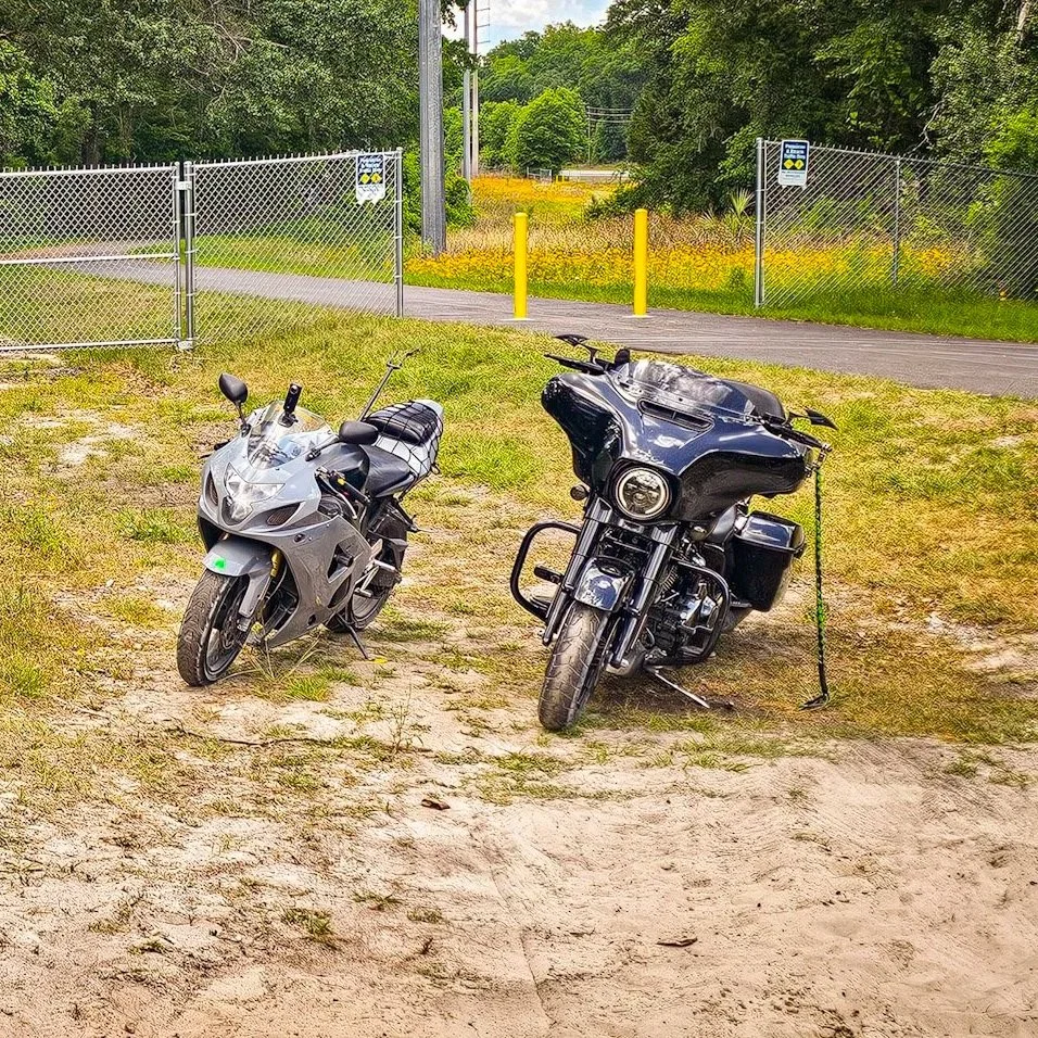 Two motorcycles parked on a grassy and dirt area near a fence with a closed gate, trees and a road in the background.