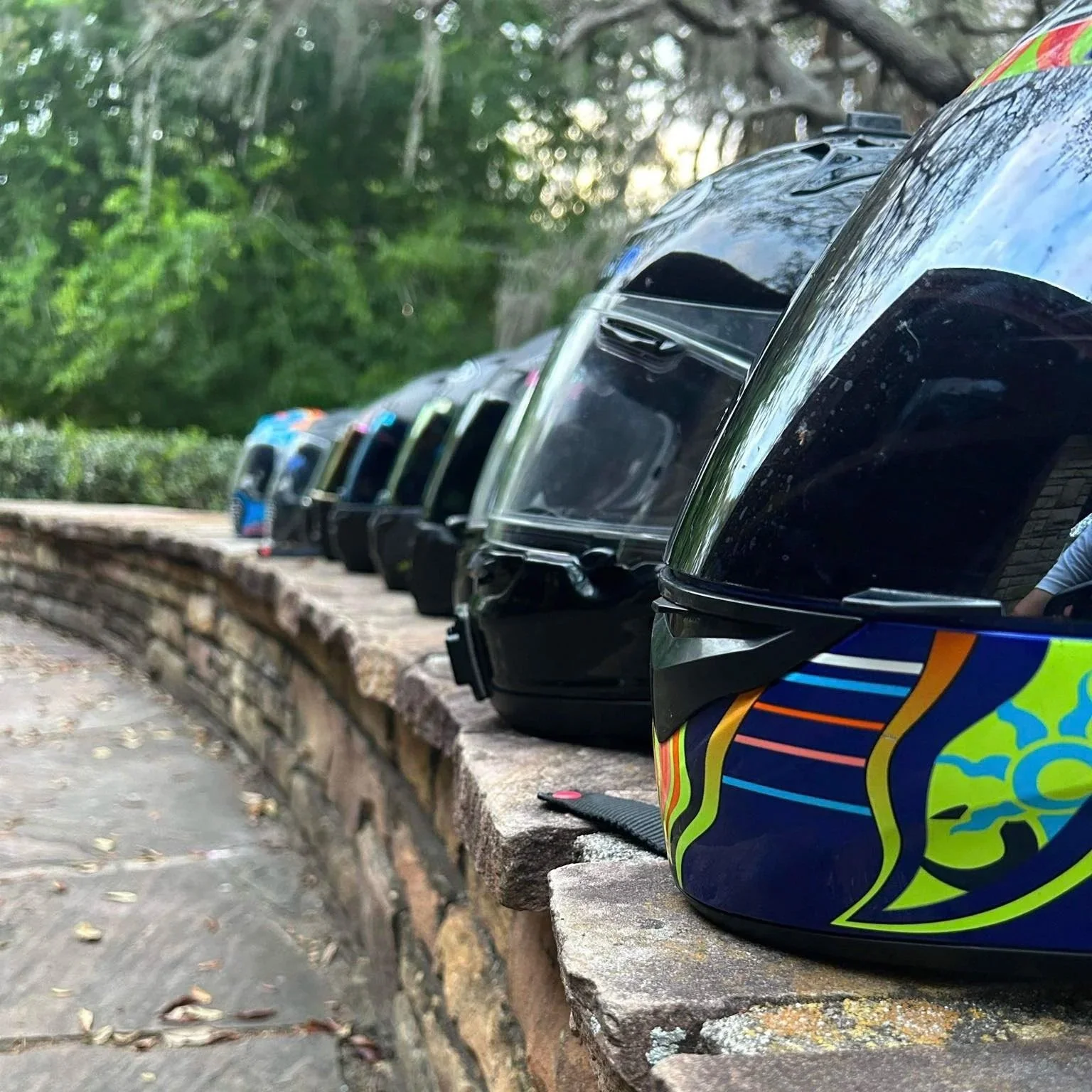 A row of motorcycle helmets lined up on a stone wall in a park with trees and greenery in the background.