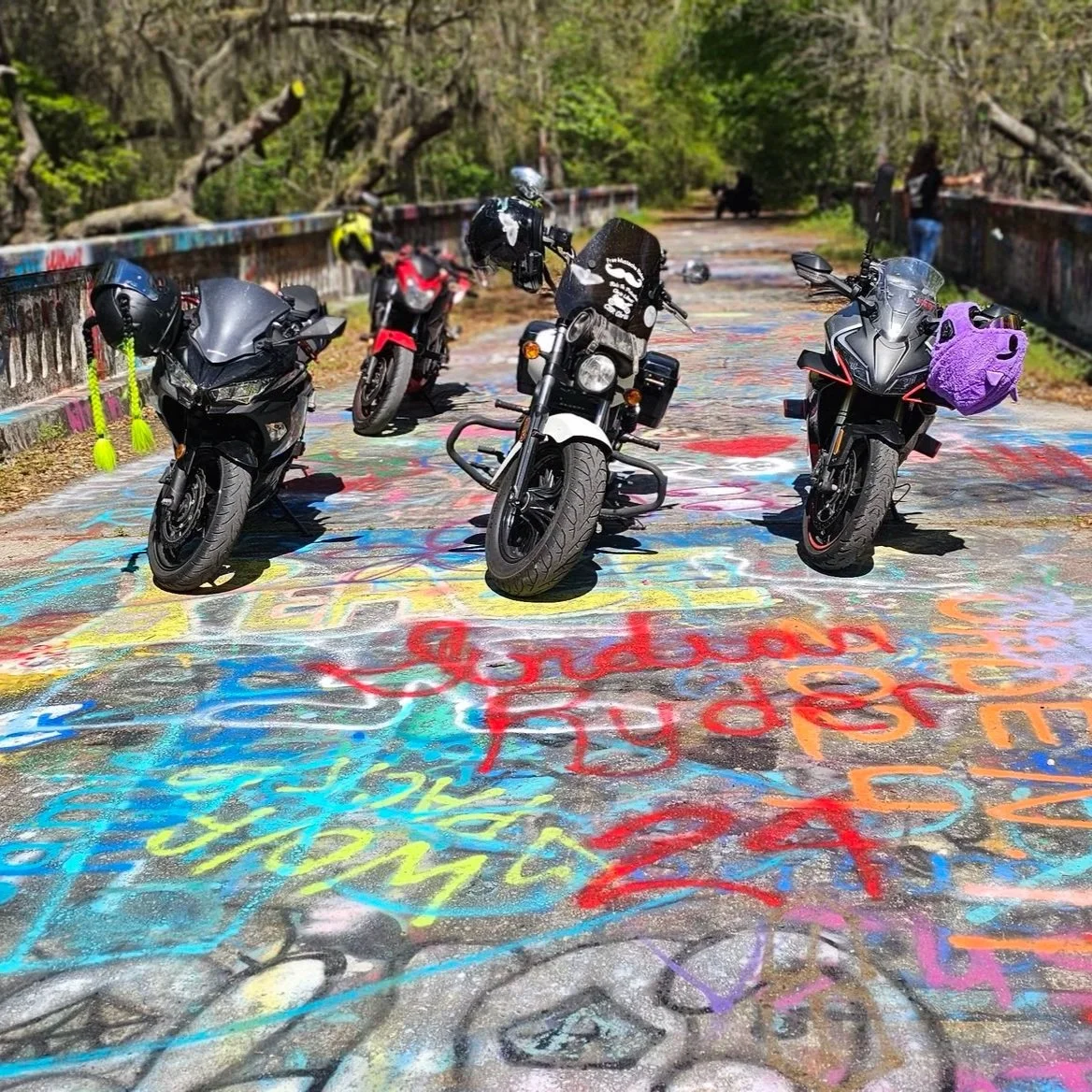 Four motorcycles parked on a graffiti-covered bridge in a wooded area.