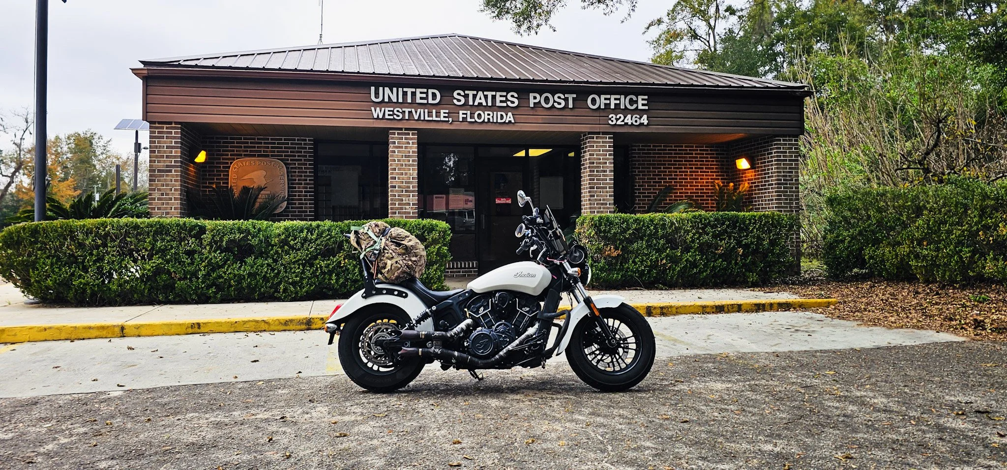 A white motorcycle parked outside a brick United States post office building in Westville, Florida, with green bushes and trees in the background.
