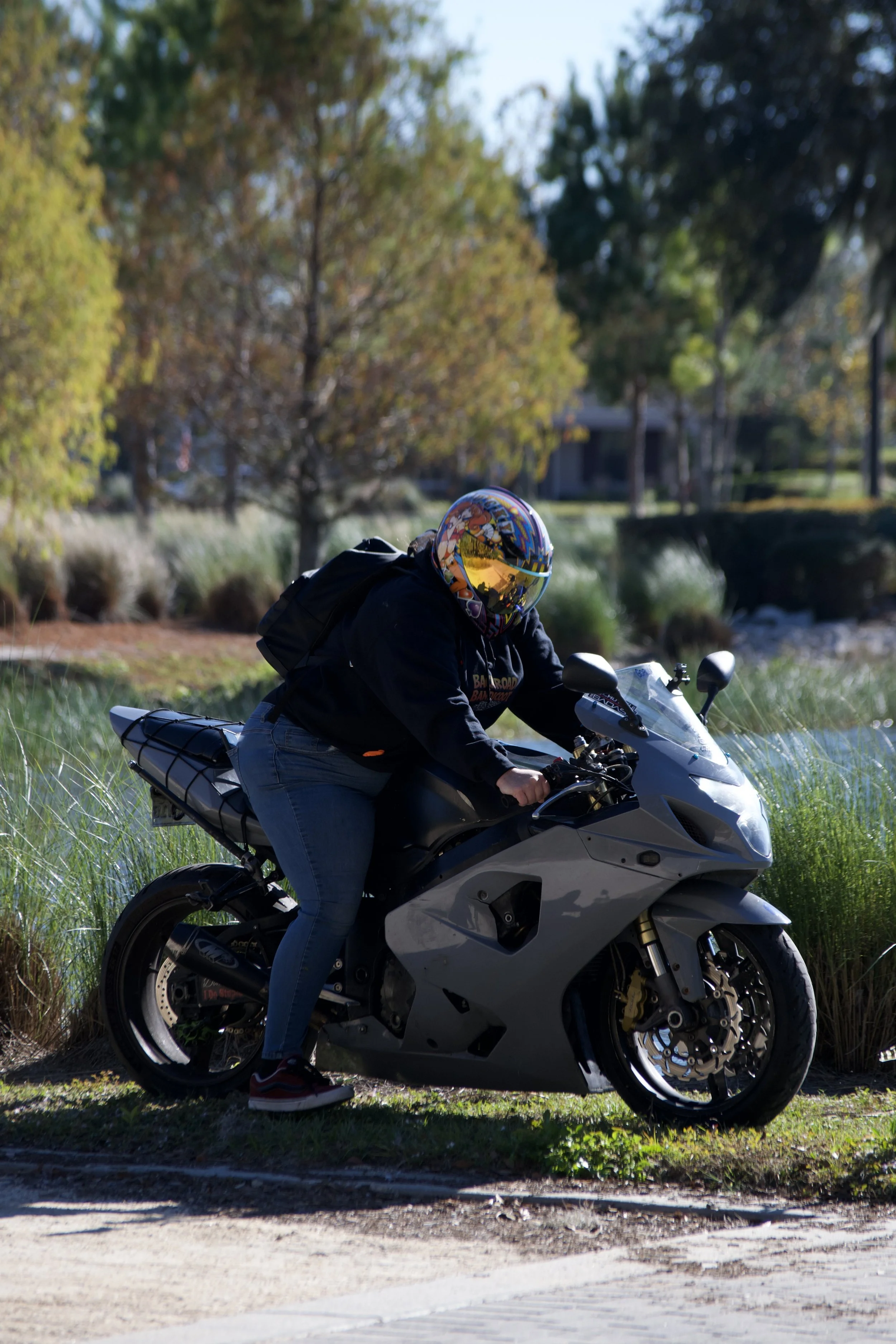 A person wearing a helmet and a black backpack is seated on a sport motorcycle on a grassy area beside a dirt path with trees and a building in the background.