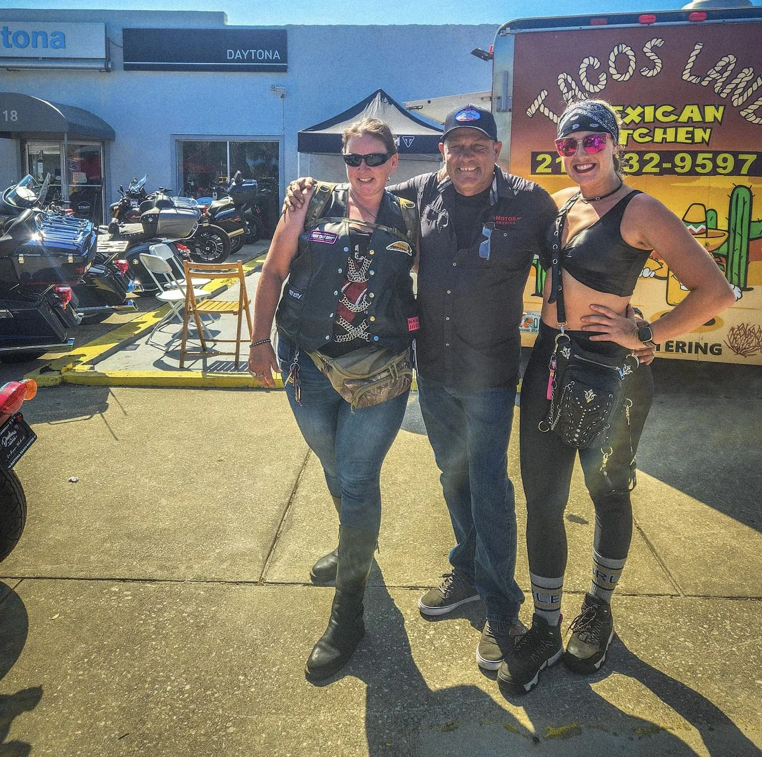 Three people standing together outdoors near motorcycles and a food truck, smiling and posing for the photo.