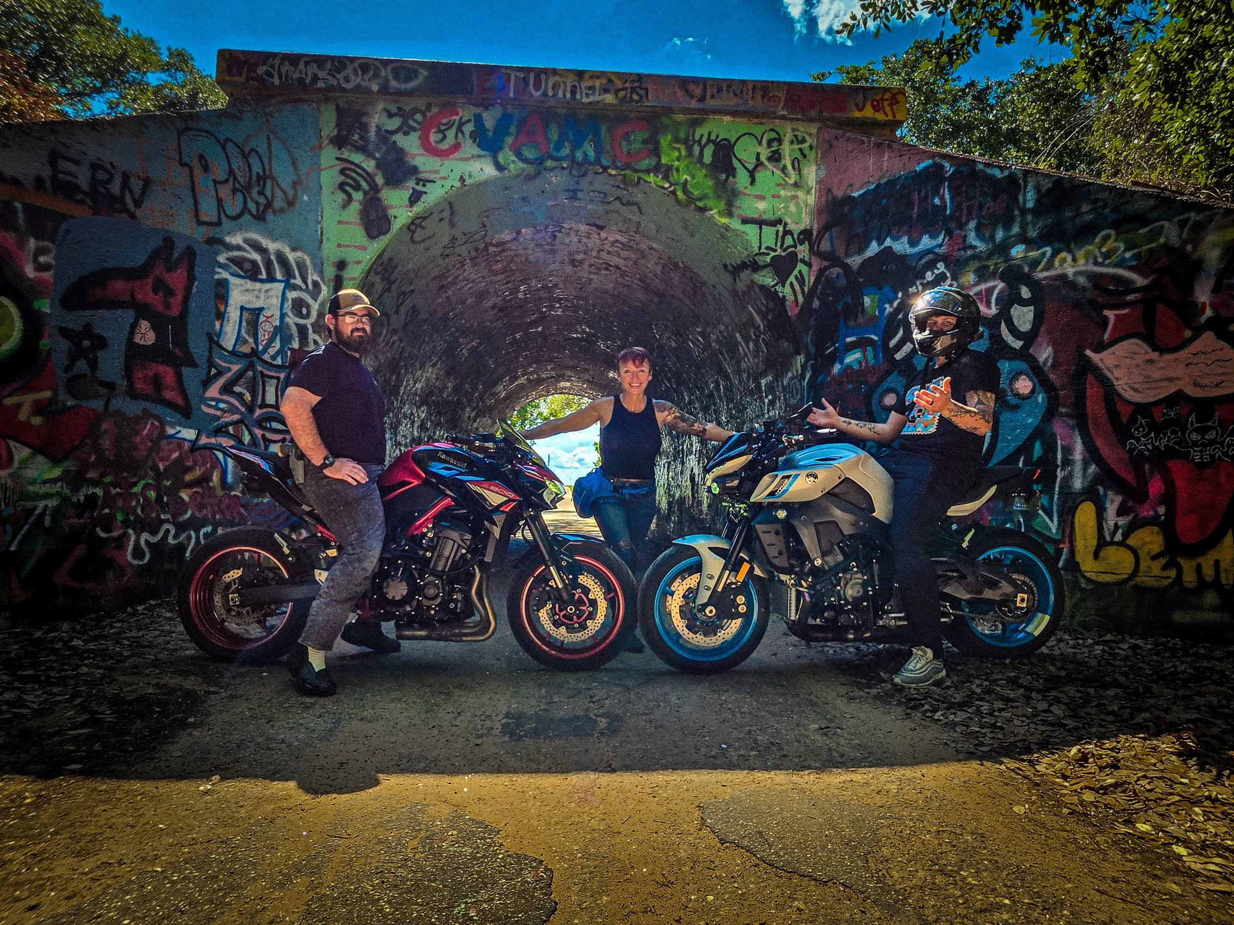 Three people on motorcycles standing under a graffiti-covered tunnel with trees in the background.