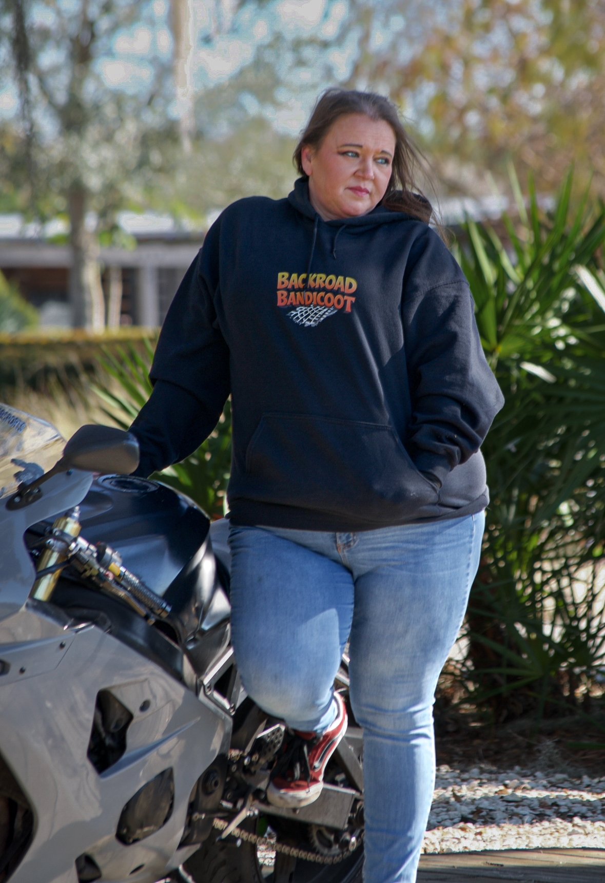A woman standing next to a motorcycle outdoors, wearing a black hoodie with 'Backroad Bandicoot' written on it and light blue jeans.