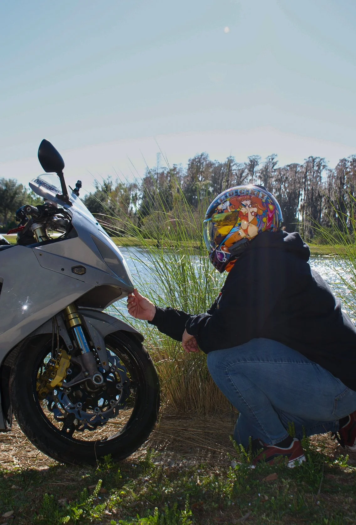 Person wearing a colorful anime-themed helmet crouching next to a silver motorcycle by a lake, touching the motorcycle's front part, with tall grass, trees, and a bright blue sky in the background.