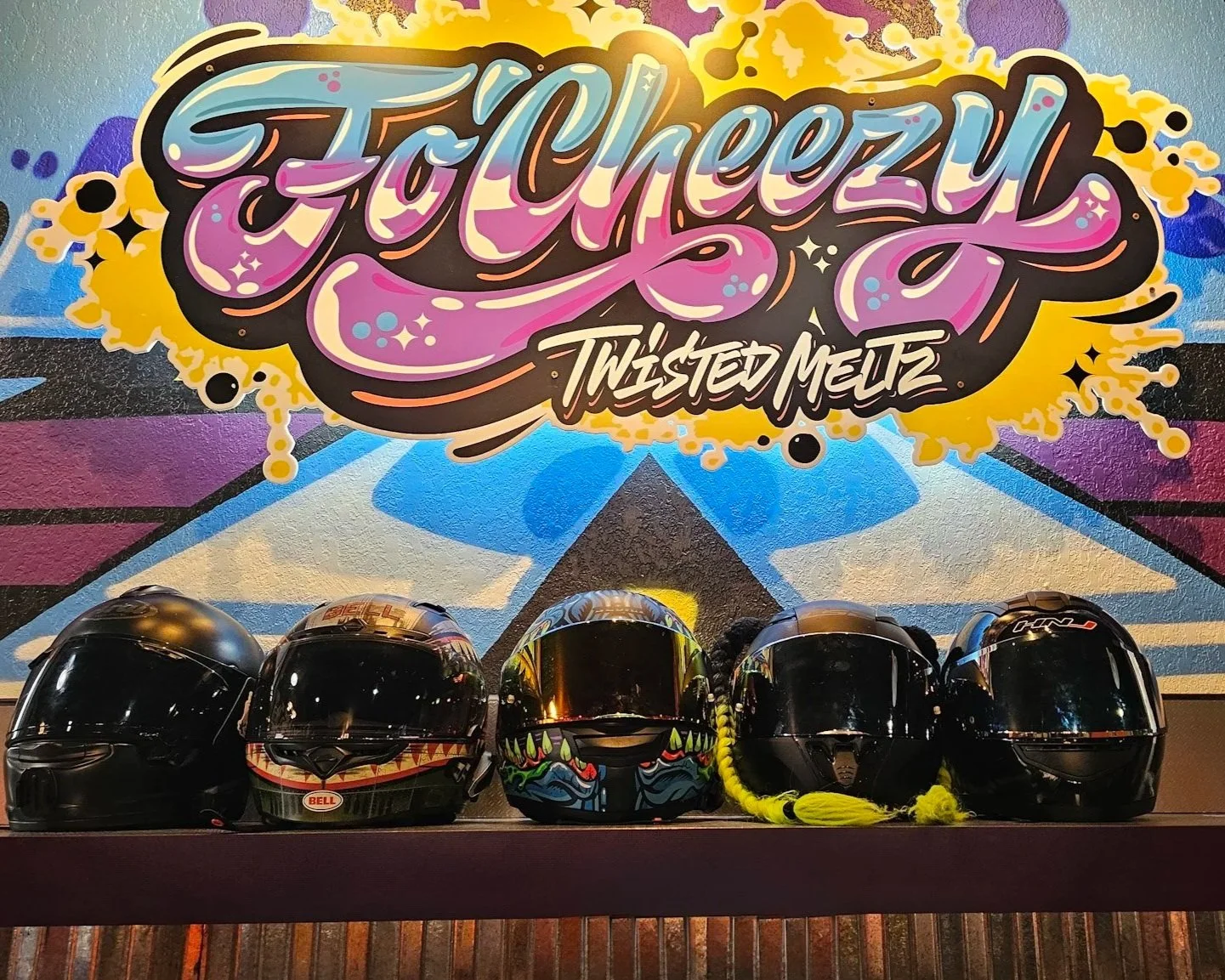 Four motorcycle helmets placed on a shelf in front of a colorful graffiti wall with the text 'Fo Cheezin' Twisted Metz' in stylized lettering.