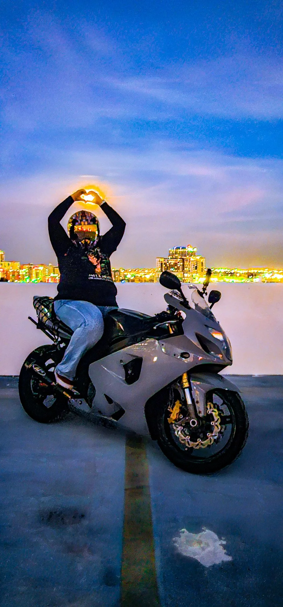 Person sitting on a sport motorcycle on a parking lot at night, making a heart shape with their hands above their head, with a cityscape and colorful sky in the background.