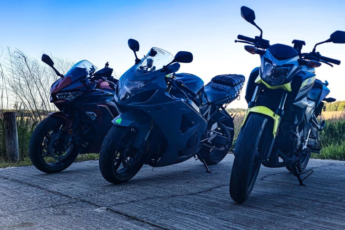 Three motorcycles parked on a concrete surface with a rural field and blue sky in the background.