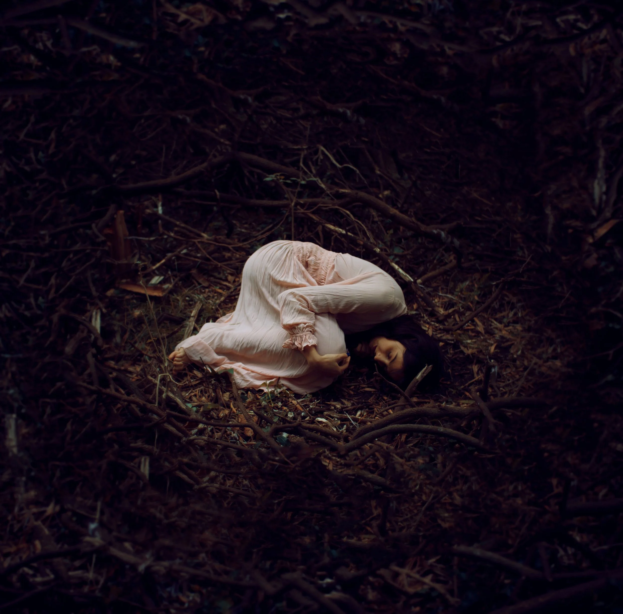 A person lying on the ground with arms extended, surrounded by dirt and bare branches or twigs. The person is wearing a light-colored garment.