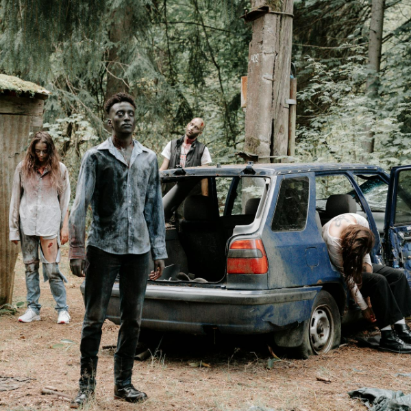 Four people in a wooded area around a blue car with an open trunk. One person is standing in the foreground, one is sitting inside the back of the car, and two others are near the car, appearing disheveled and distressed.