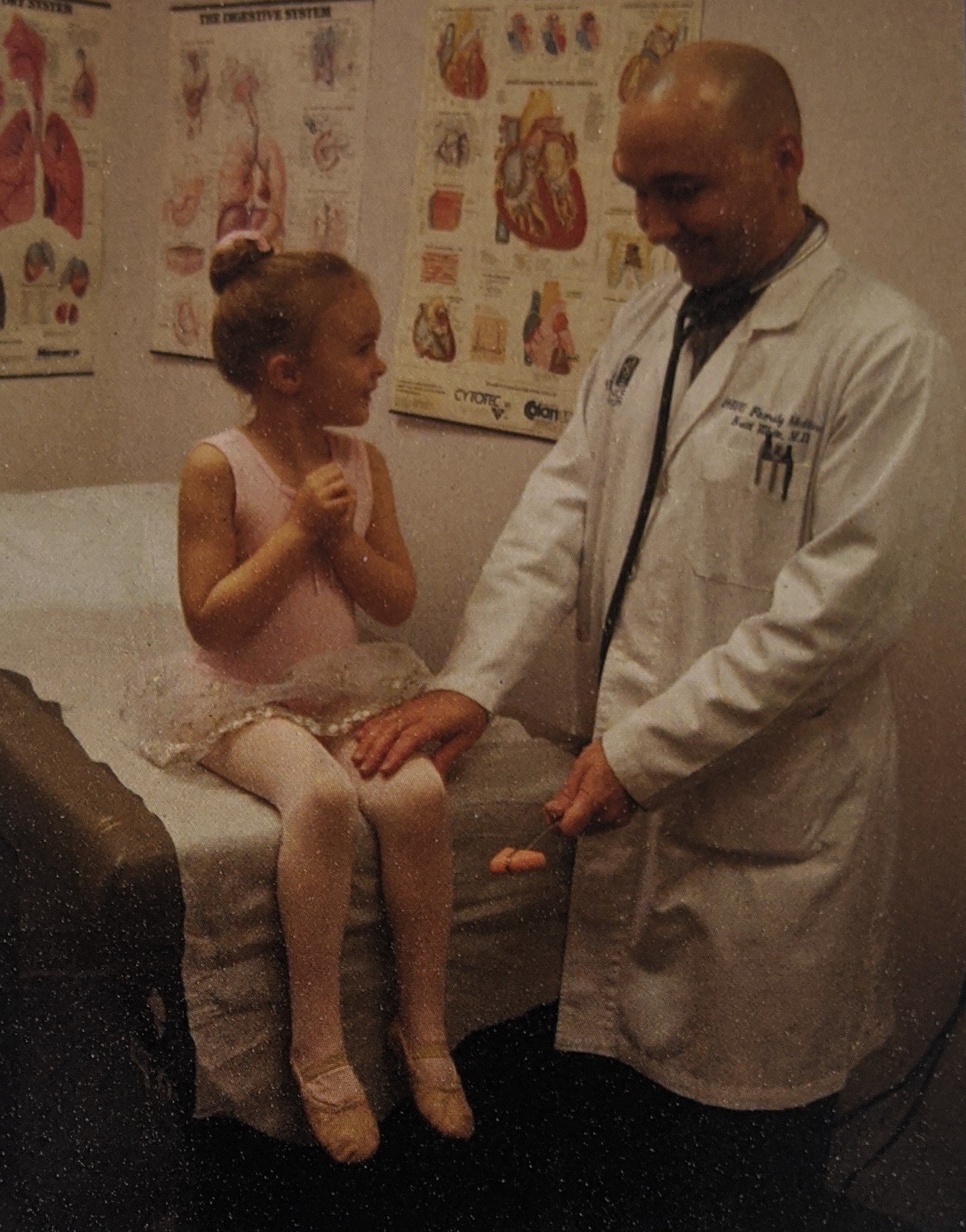 A young girl in ballet attire sitting on an examination table in a medical office, talking to a male doctor in a white coat, who is holding a reflex hammer. Posters of human anatomy are on the wall behind them.