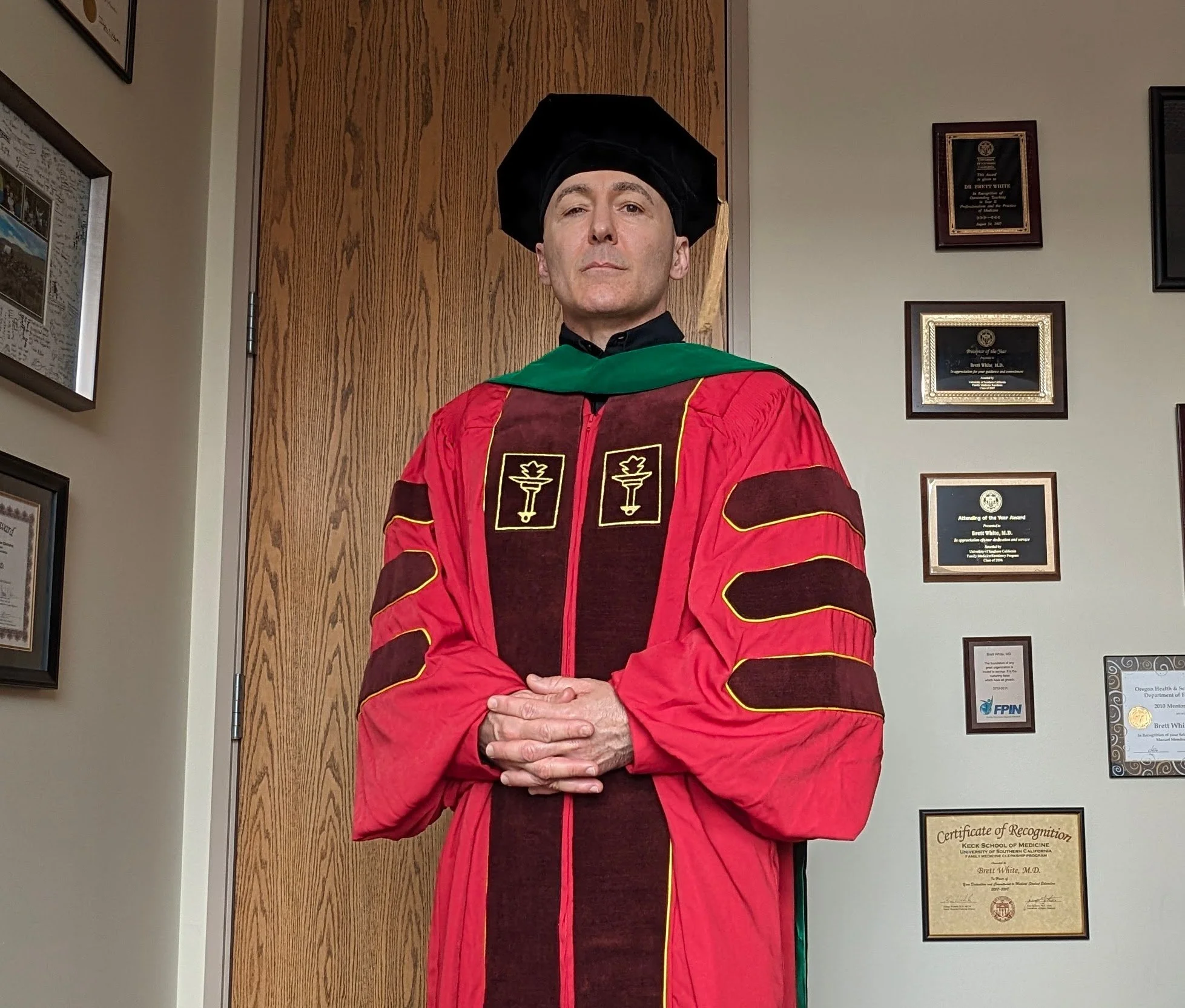 A man dressed in academic regalia, including a black cap and a red gown with gold piping, standing with hands clasped in front of a wooden door and framed awards and certificates on the wall.