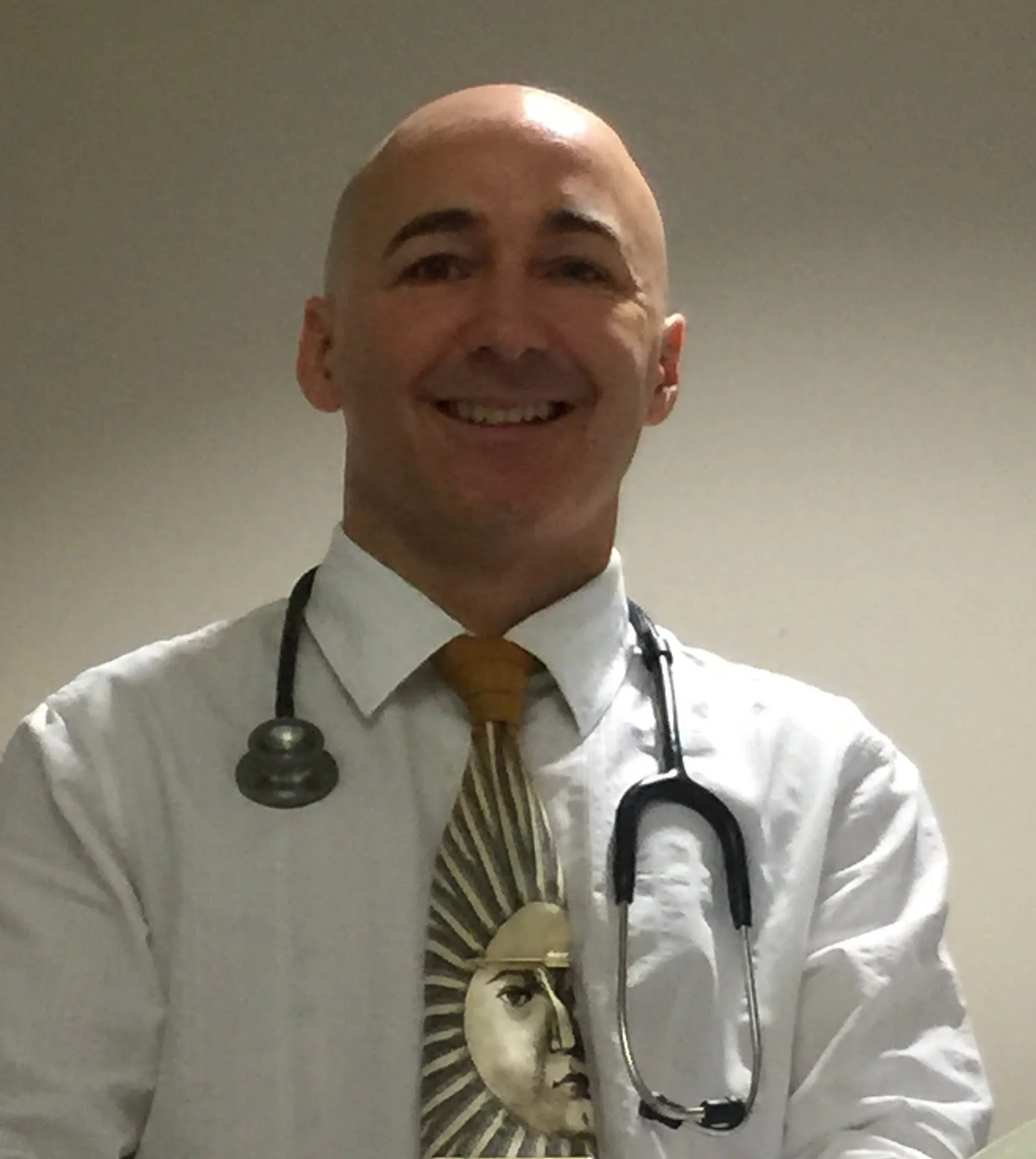 A man wearing a white coat with a stethoscope around his neck, smiling, with a shirt featuring a portrait of a woman and a striped tie.
