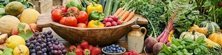 A large wooden bowl filled with various fresh fruits and vegetables, including tomatoes, bell peppers, cucumbers, carrots, and radishes, surrounded by more fruits and vegetables like grapes, strawberries, beets, lettuce, and squash on a wooden table.