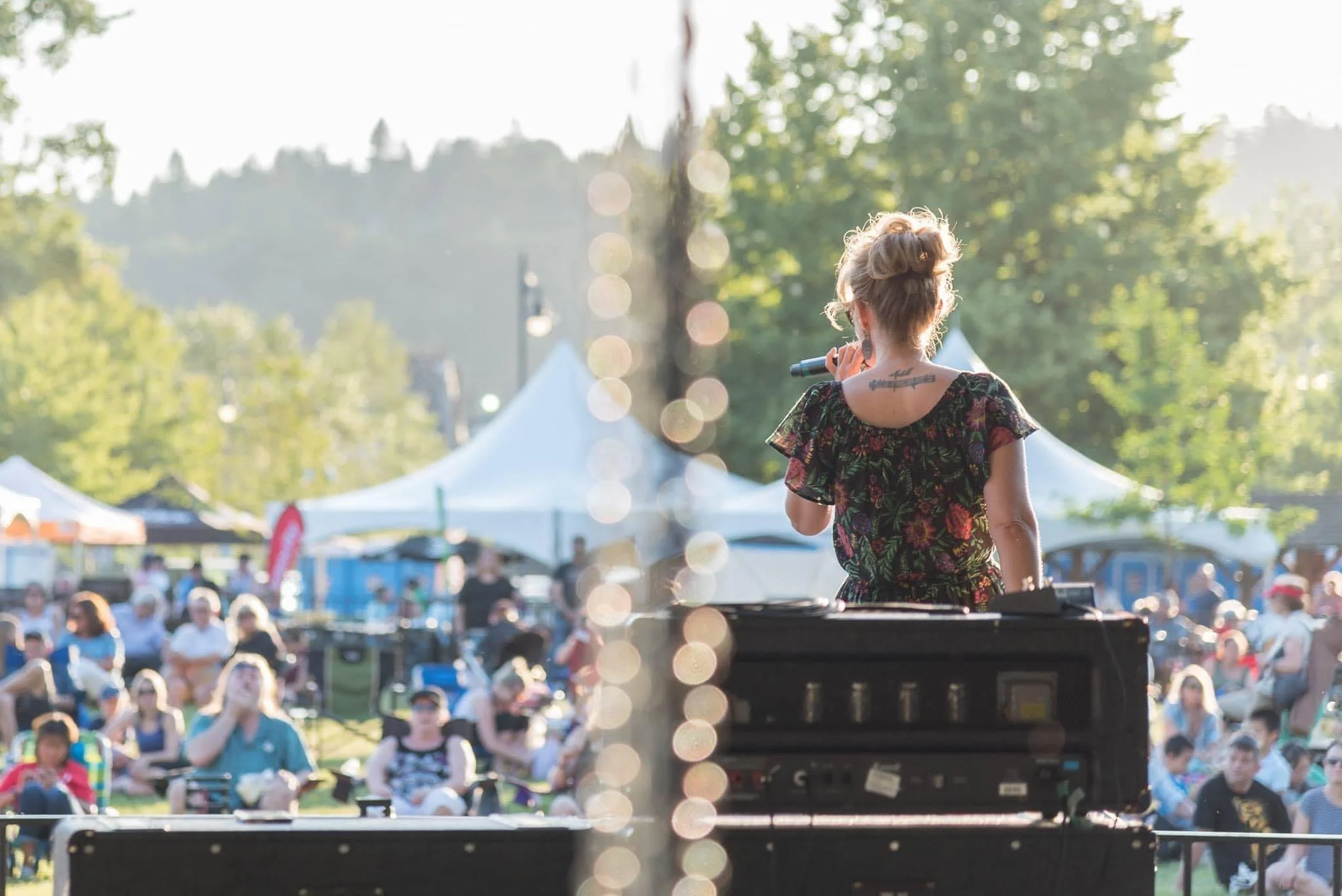 A woman with blonde hair in a bun, wearing a floral dress, standing in front of a crowd at an outdoor event, holding a microphone.