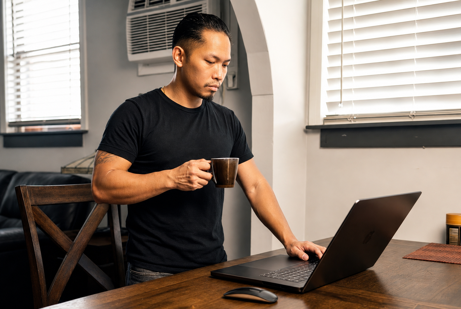 A man holds a coffee mug while working on a laptop at a wooden table in a room with white blinds on the window.