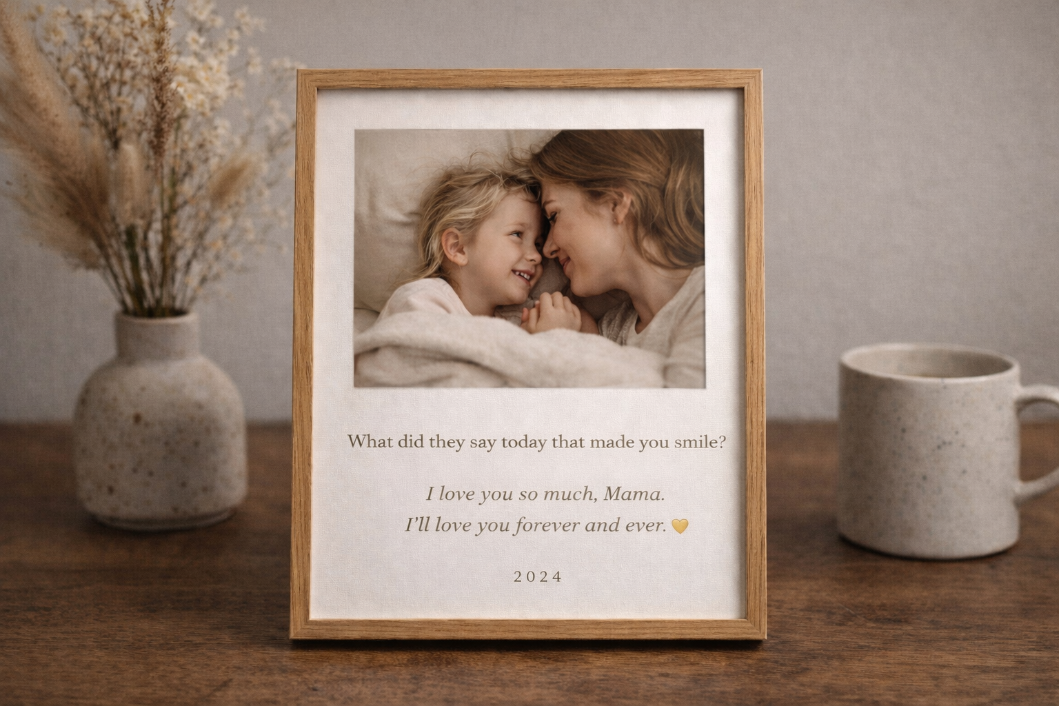 A framed photo of a mother and daughter smiling and touching foreheads while lying in bed, with a heartfelt message below about their love, surrounded by decorative vases and dried flowers on a wooden surface.