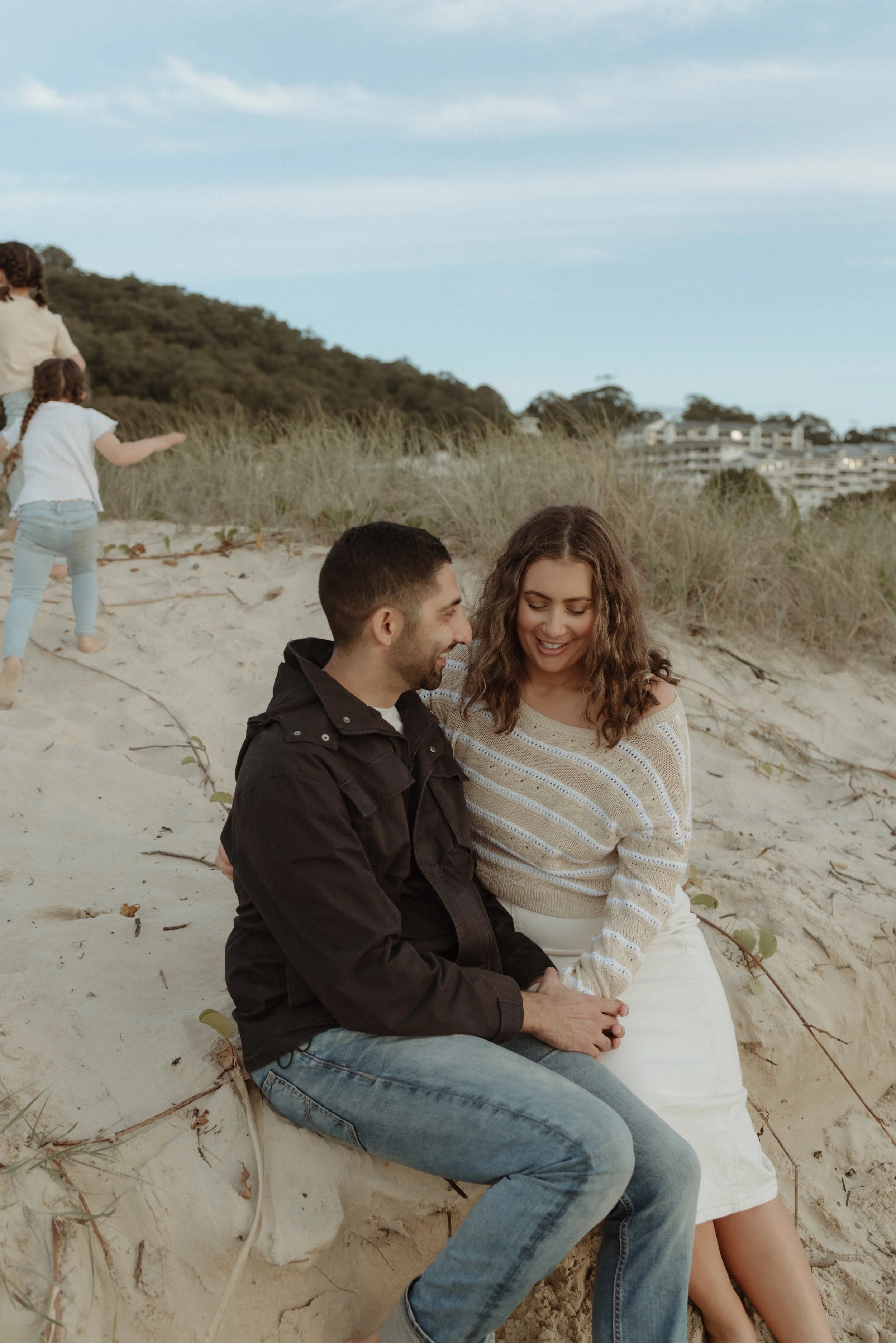 A young couple sitting close together on the sandy beach, smiling and looking at each other, with children running in the background near grassy dunes under a blue sky.
