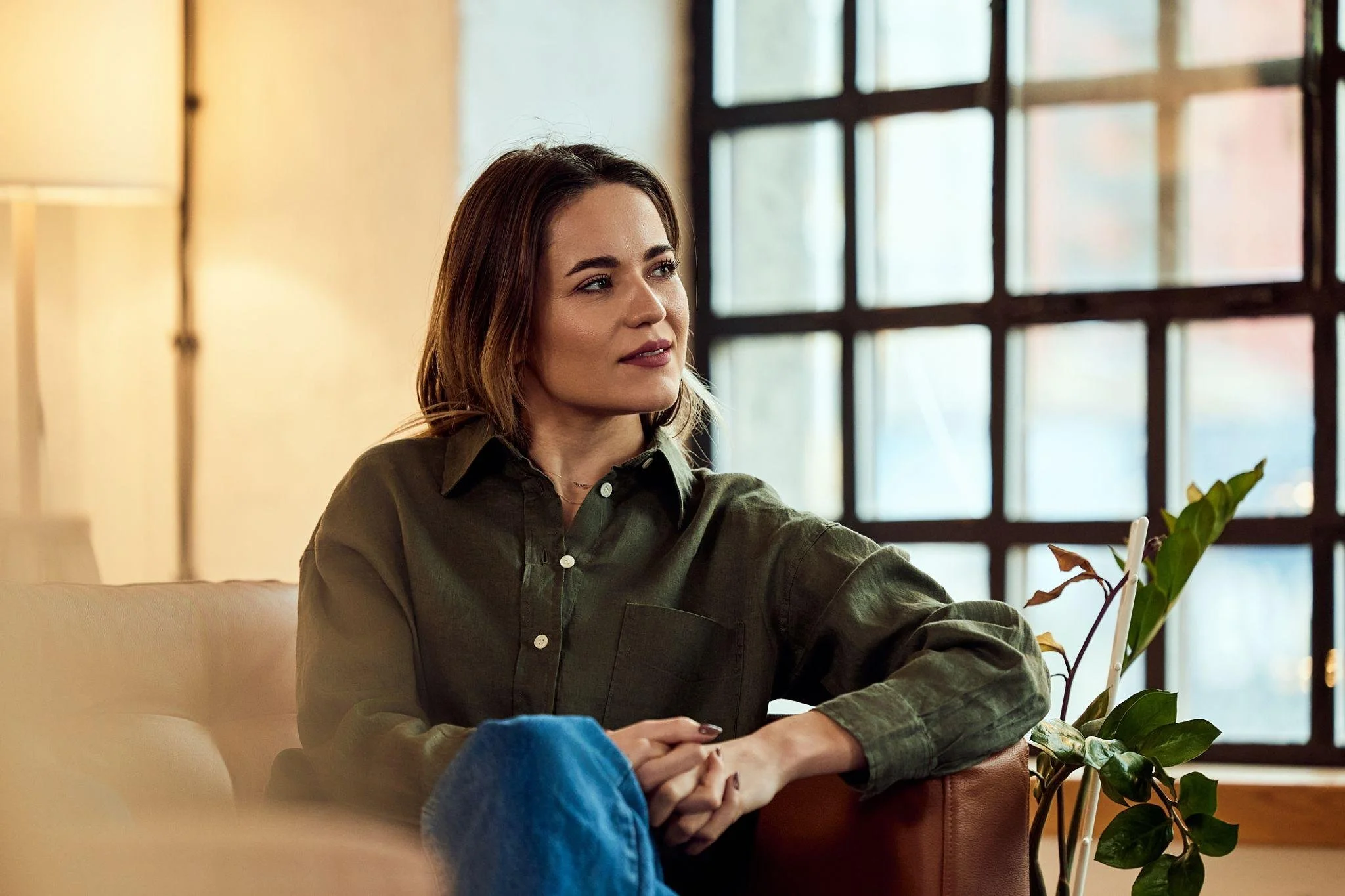 A woman with shoulder-length brown hair and a green shirt sitting at a table near a window with large panes, next to some potted plants.