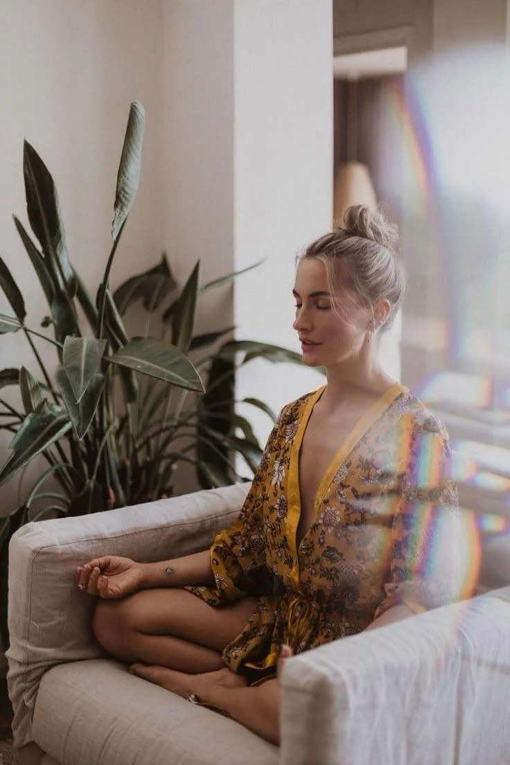 A woman with blonde hair in a bun practicing meditation indoors, sitting cross-legged on a cream-colored couch near a large leafy plant, wearing a yellow floral robe with closed eyes and a peaceful expression, with sunlight creating rainbow light refractions.
