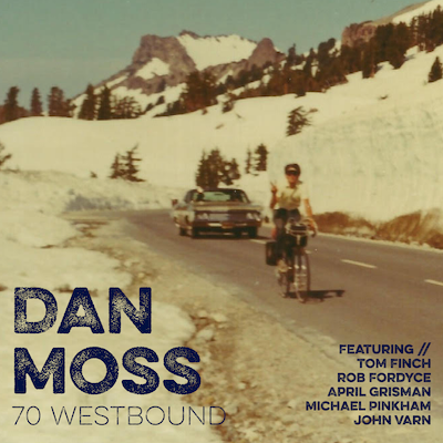 A person riding a bicycle on a snowy mountain road, with snow-covered mountains and trees in the background, and a car on the road, with text promoting Dan Moss and other performers.