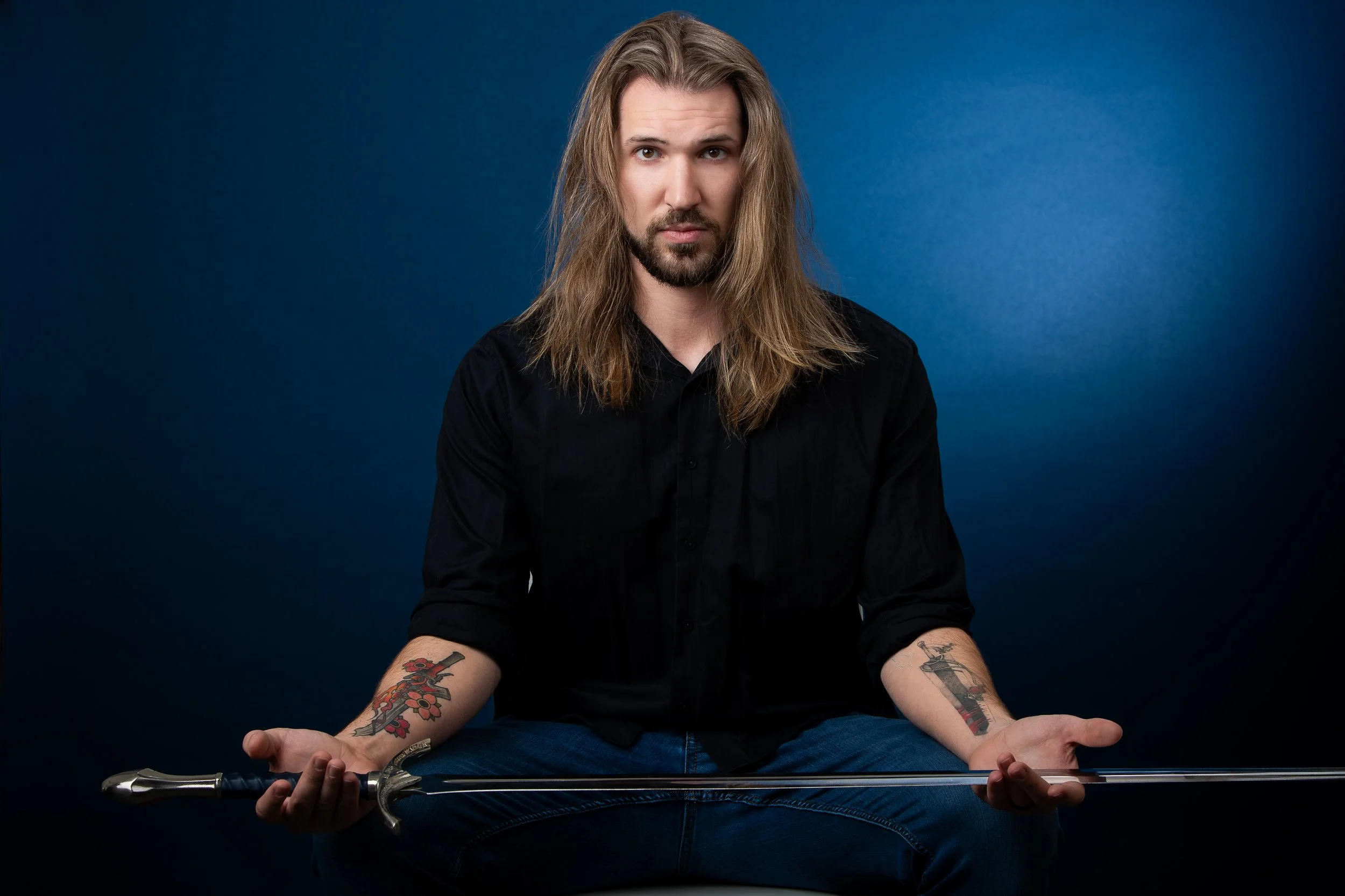 A man with long hair, a beard, and tattoos on his arms, dressed in a black shirt, sits cross-legged against a blue background, holding a sword.