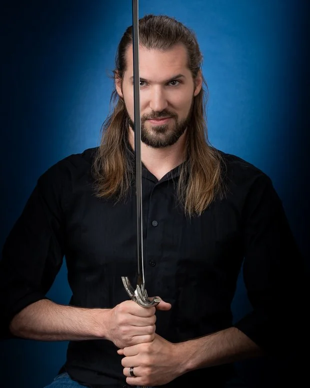 Man with long hair and beard holding a sword vertically in front of his face, wearing a black shirt, against a blue background.
