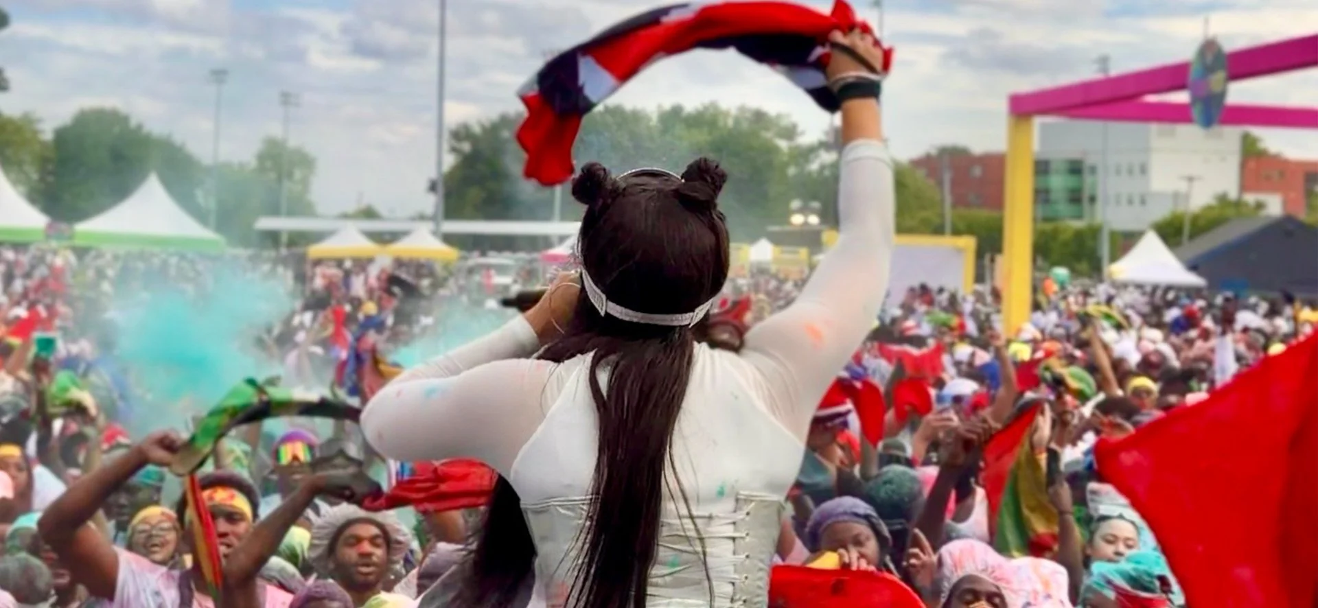 Jadel with long dark hair styled in two buns, wearing a white long-sleeve shirt, stands with her back to the camera at a lively outdoor festival. She holds a red, white, and blue cloth in the air. Mess Fete New York 2026